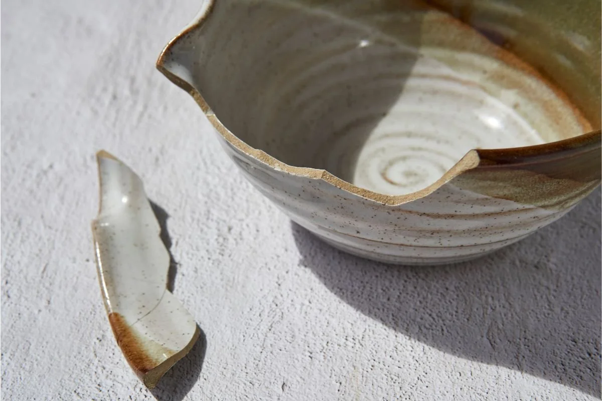 A close-up of a broken ceramic bowl on a textured white surface with a large shard lying next to it, symbolizing a lack of boundaries and energy leakage in burnout.