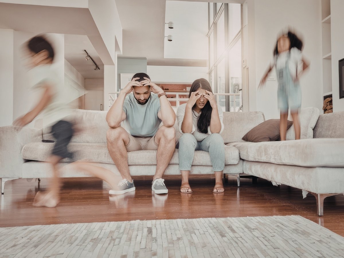 Overwhelmed parents sitting head-in-hands on a living room sofa while young children run past in motion blur.