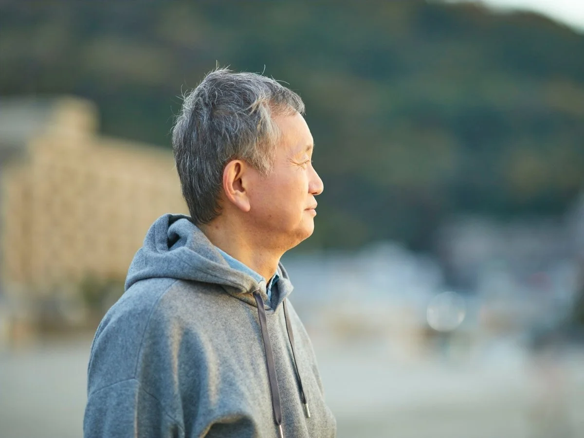 Mature man with grey hair in a casual hoodie, looking out toward the horizon in quiet contemplation outdoors.