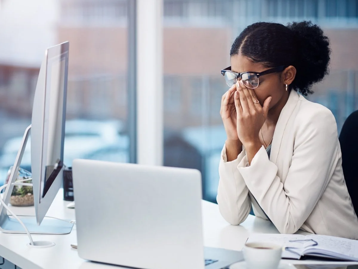 Professional executive sitting at a desk with eyes closed, pinching the bridge of her nose in deep fatigue in front of her laptop.