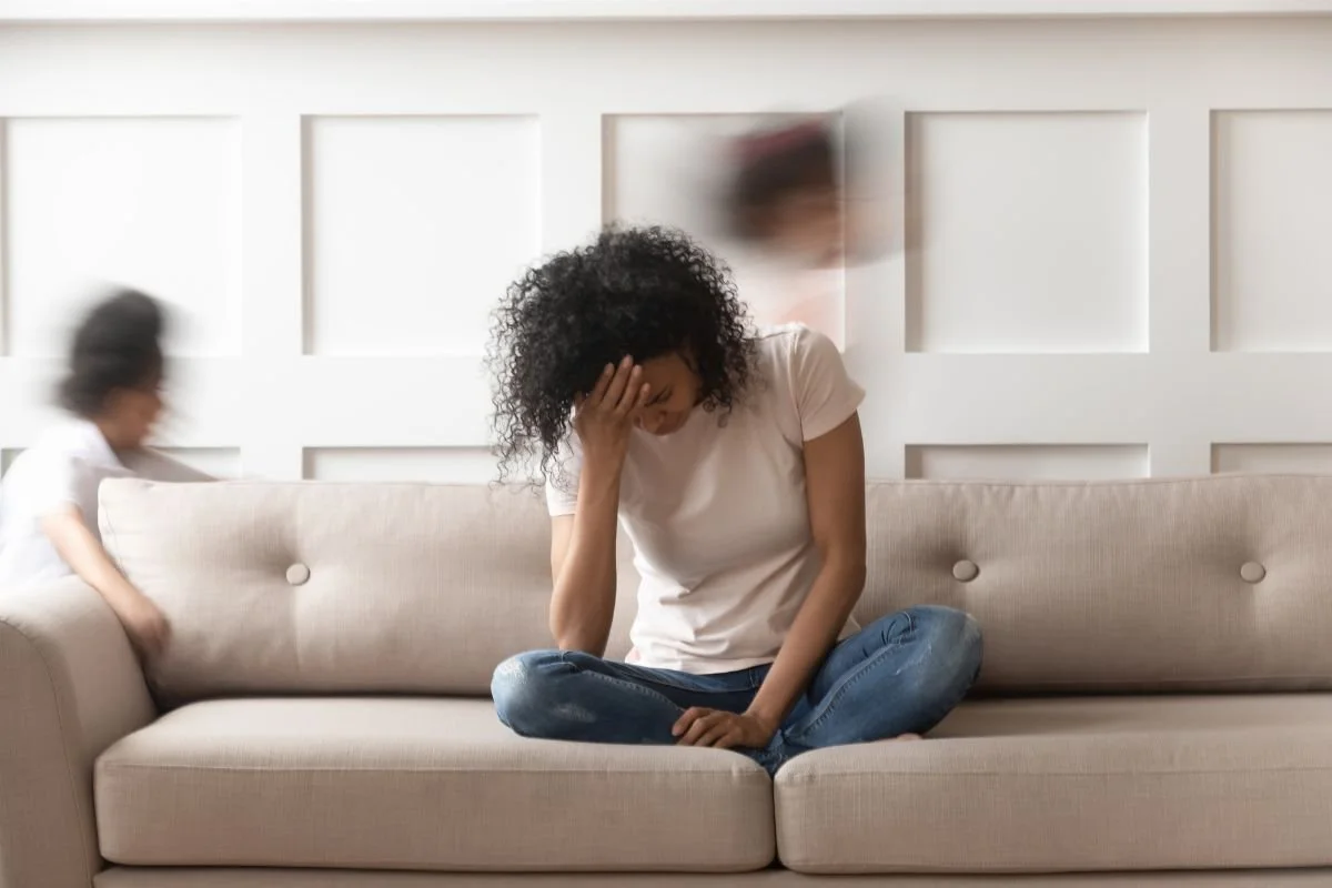 A woman with curly hair sits on a beige sofa, head in her hand, looking overwhelmed and exhausted. Blurred figures of children moving around her create a sense of chaos, illustrating the physical and emotional weight of burnout and chronic fatigue.
