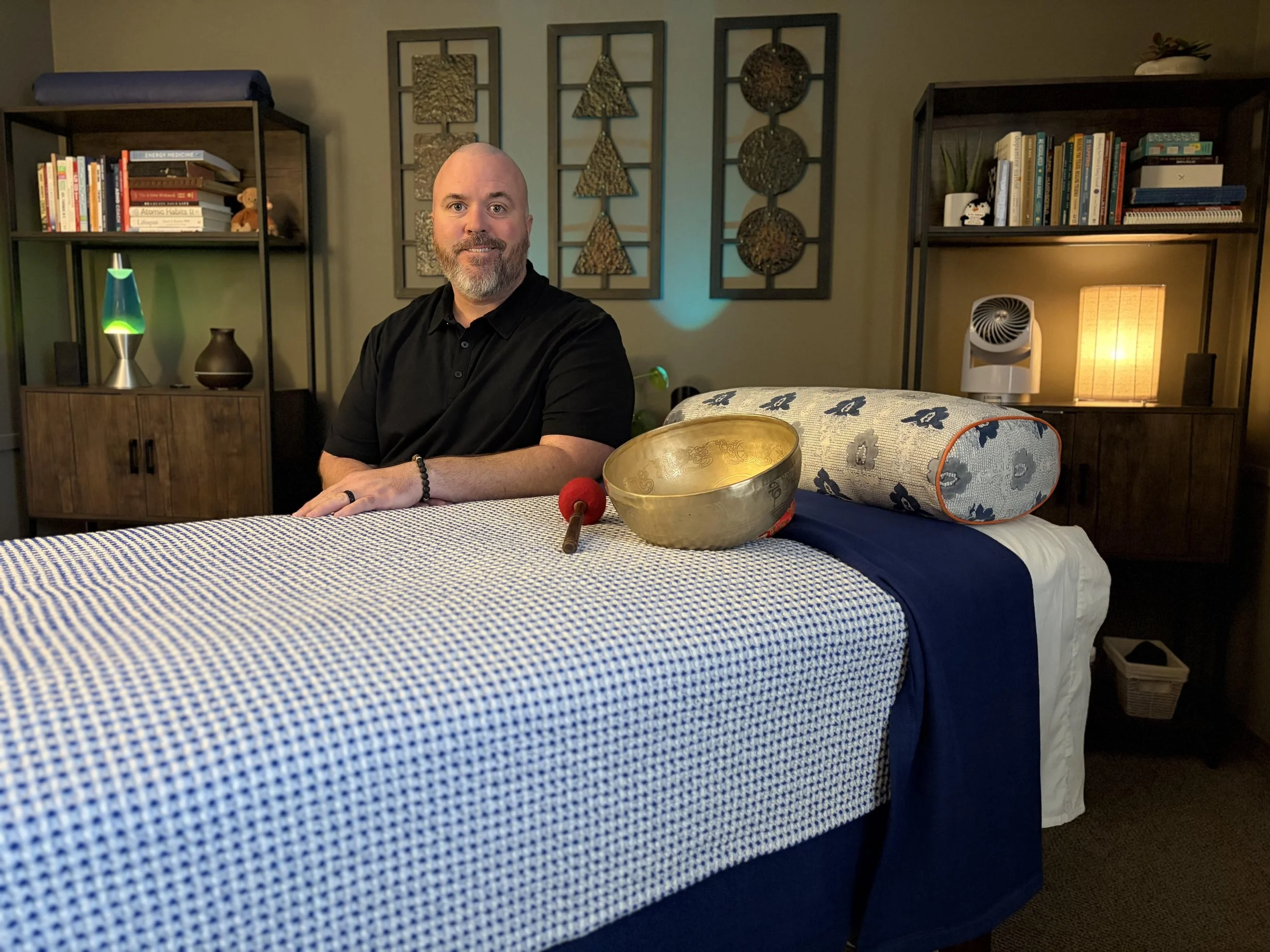 The reSpark KC practitioner seated next to a treatment table and golden singing bowl in his Overland Park integrative healing and Reiki clinic.