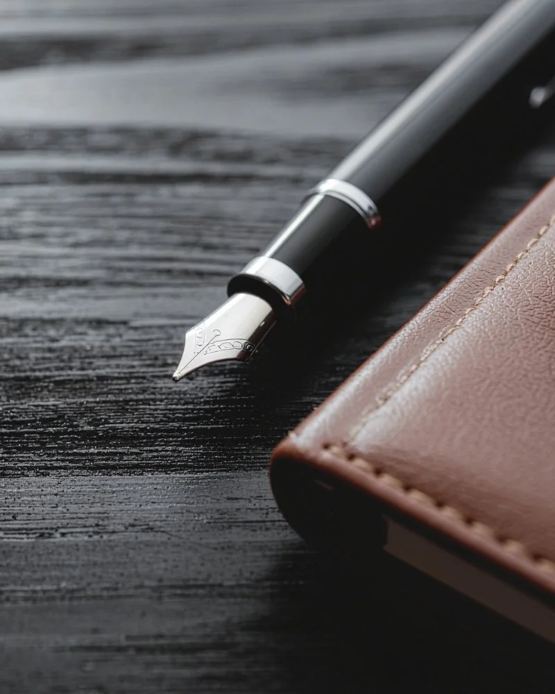 Close-up macro photograph of a sleek black fountain pen with an intricate silver nib resting on a dark, textured black wooden desktop next to the corner of a brown stitched leather journal.
