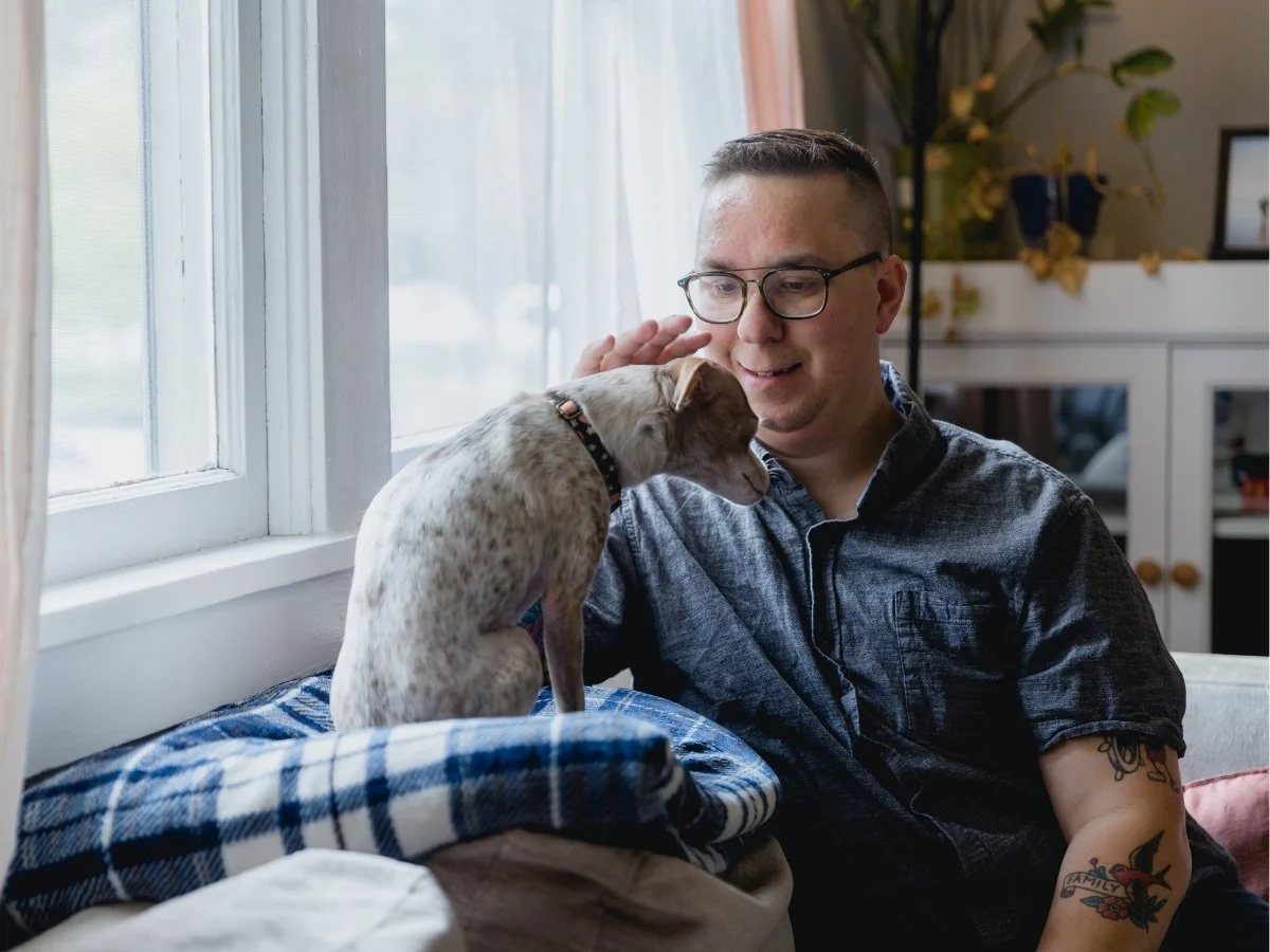 Person wearing glasses sitting by a bright window, gently petting a small dog for quiet somatic grounding.
