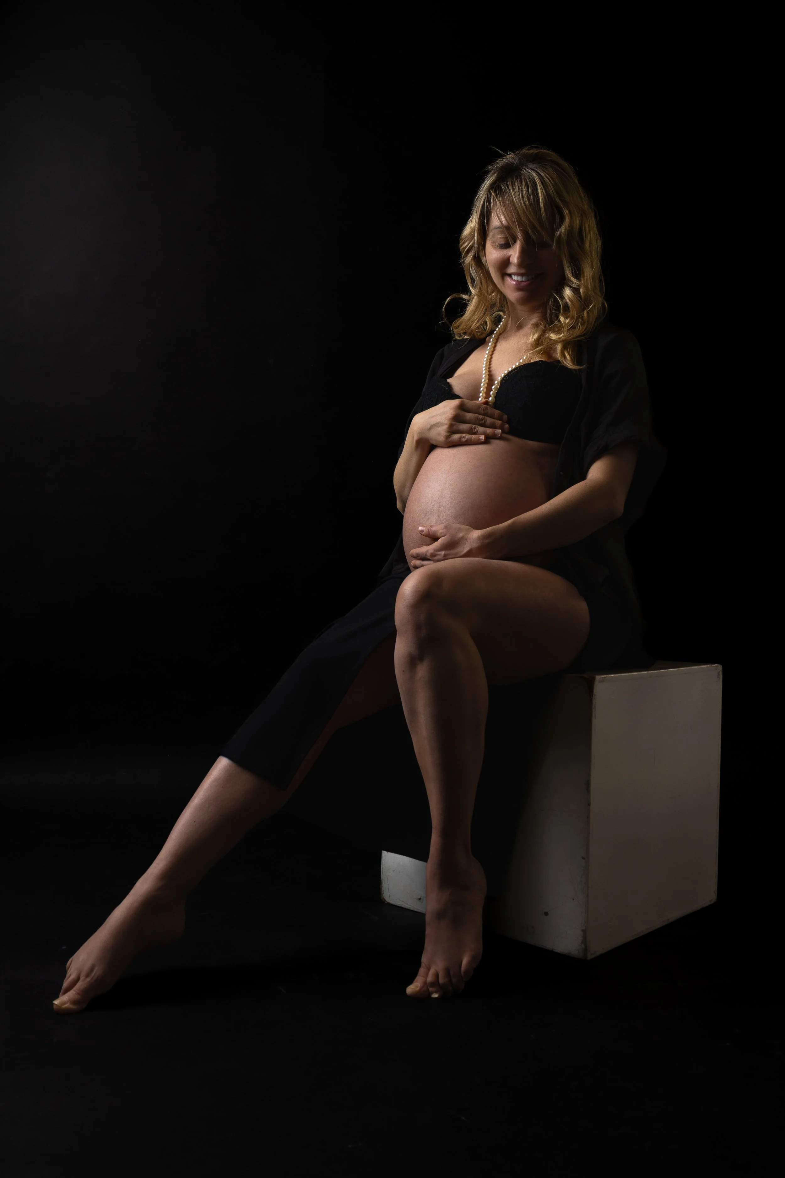 Pregnant woman sitting on a white block, gently holding her belly, with a black background.