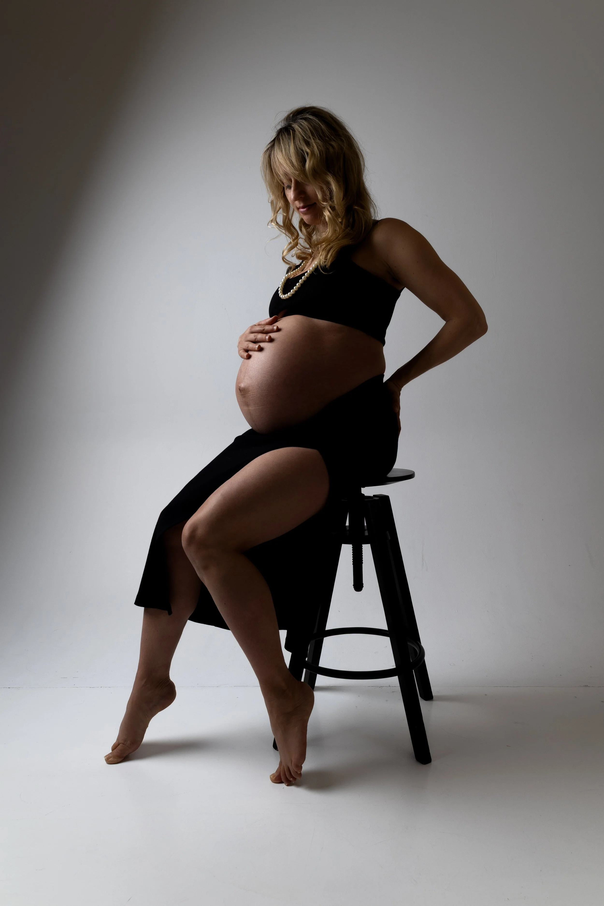 A pregnant woman with curly blonde hair sitting on a black stool against a plain white background, wearing black clothing and a pearl necklace.