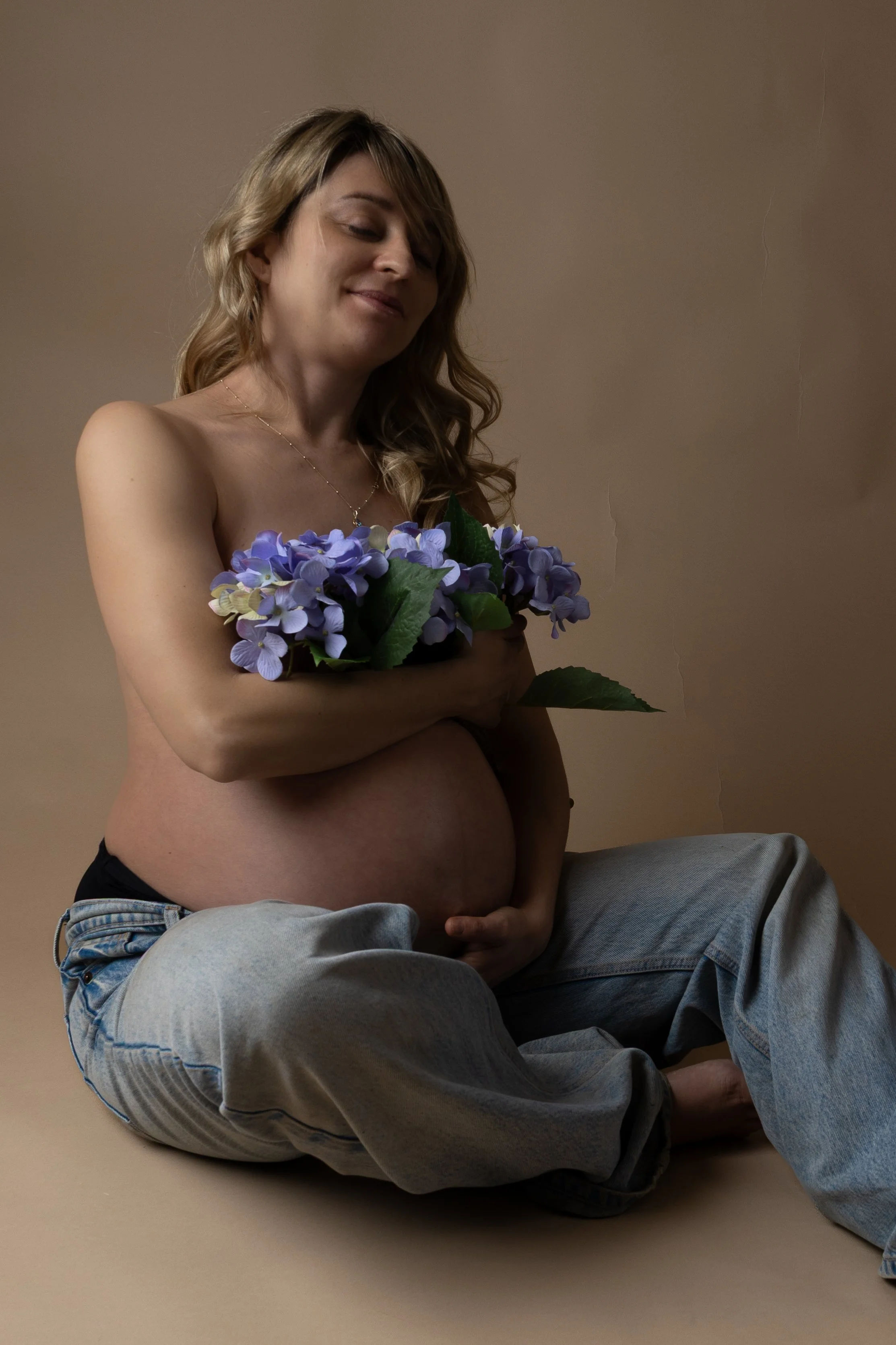 A pregnant woman sitting cross-legged against a plain background, holding a bouquet of purple hydrangeas, smiling softly with eyes closed.