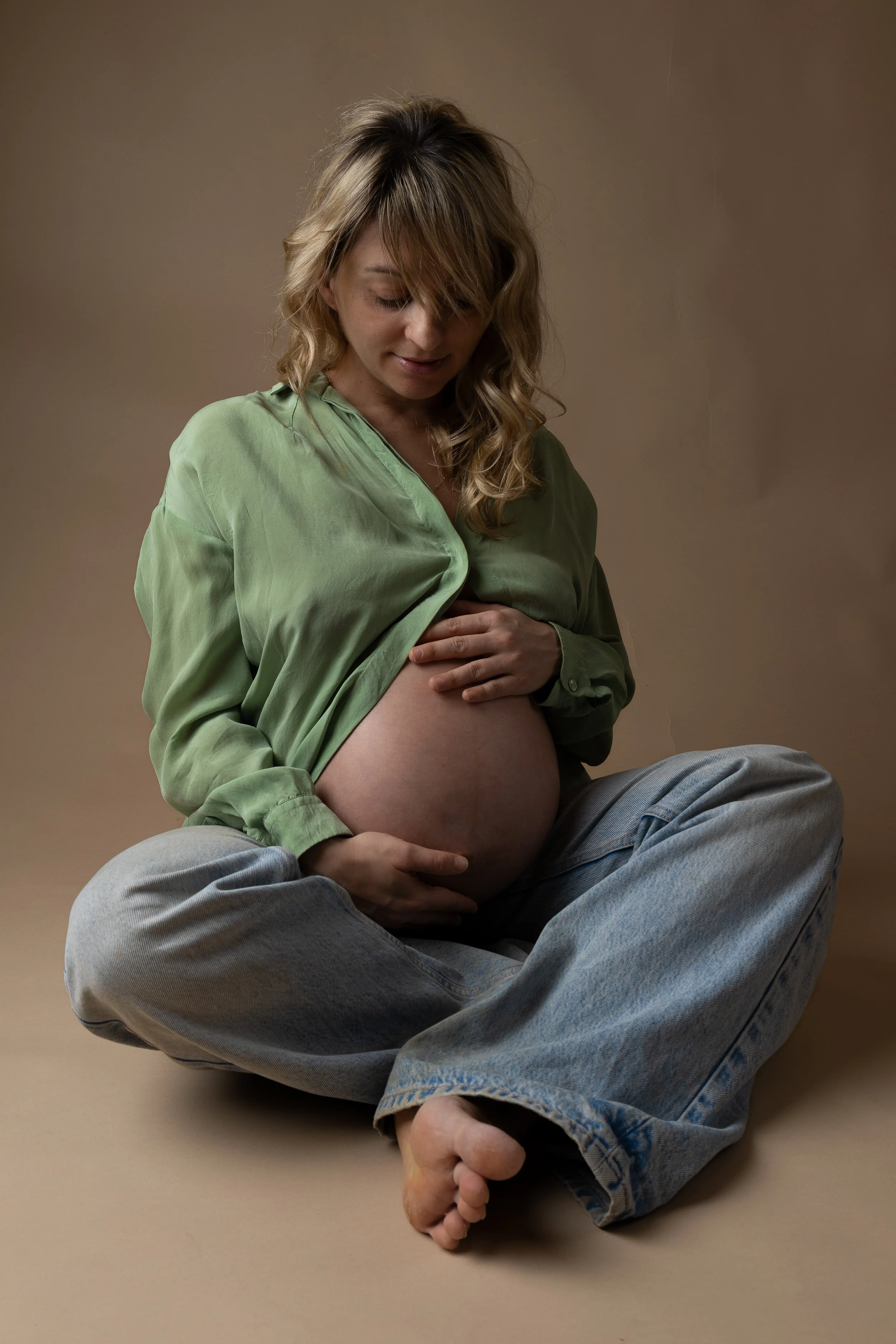 Pregnant woman sitting cross-legged on the floor, wearing a green shirt and jeans, touching her belly with both hands, looking down.