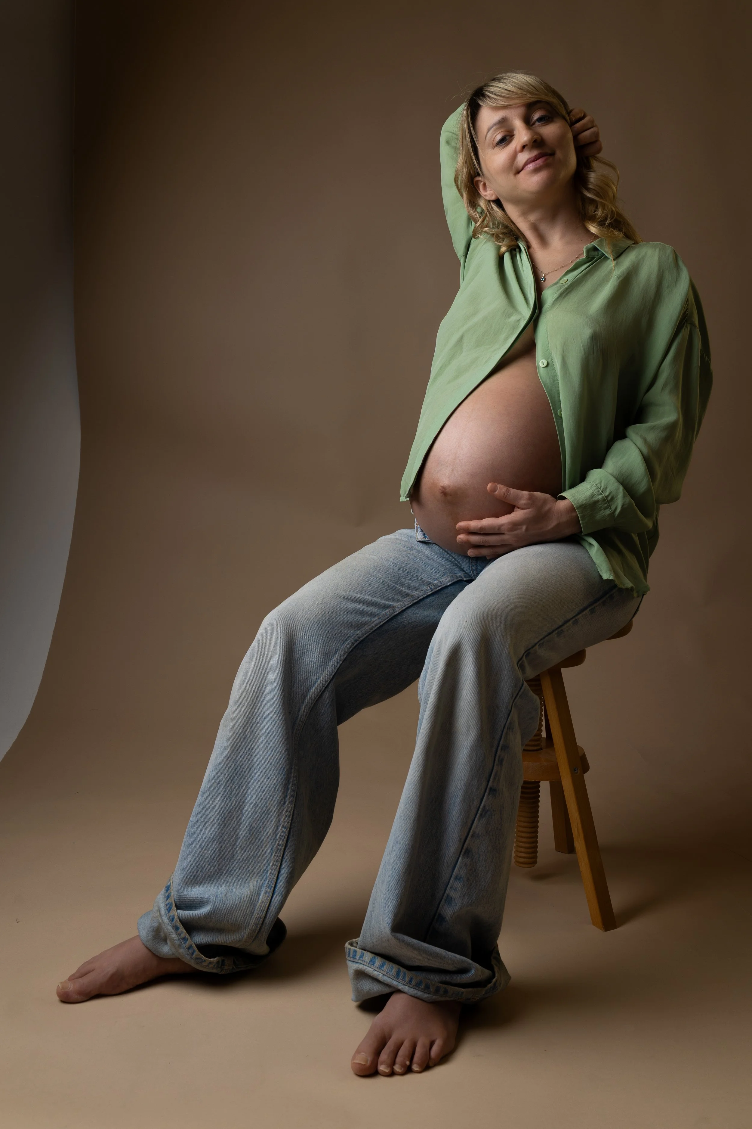 A pregnant woman sitting on a stool, wearing an open green shirt and relaxed jeans, with one hand resting on her belly and the other behind her head, looking at the camera with a gentle expression.