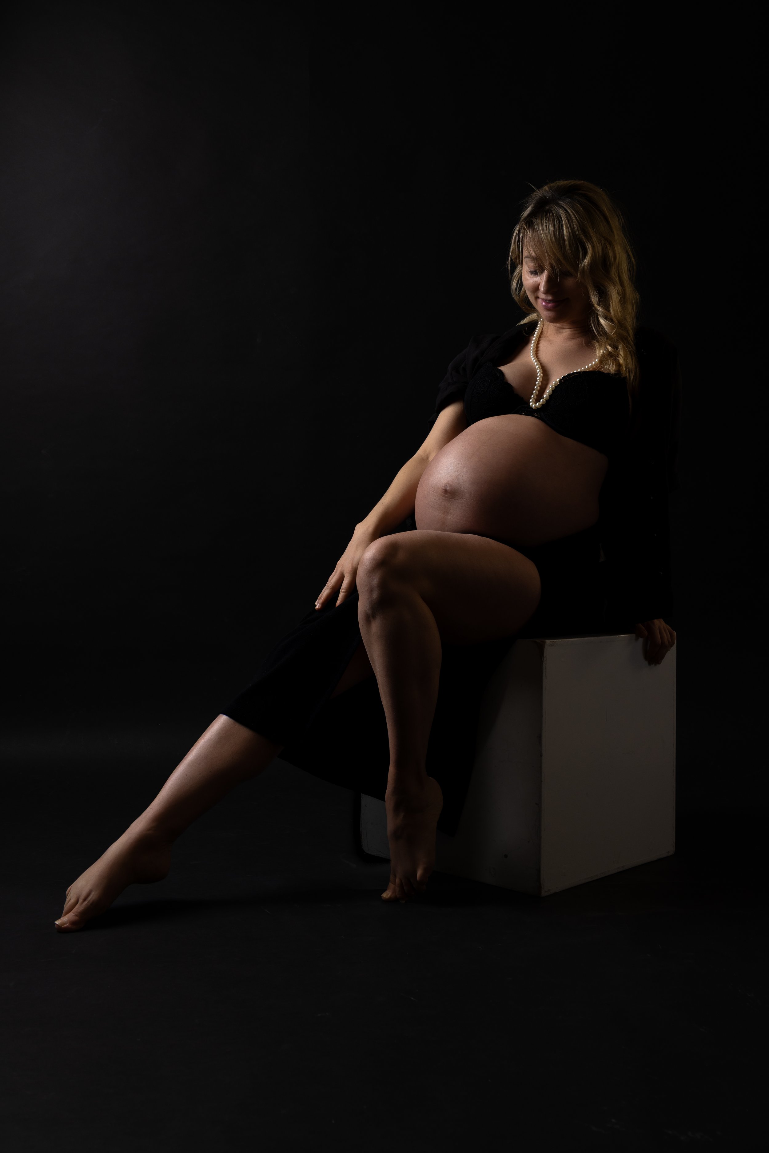 Pregnant woman sitting on a white cube, wearing a black dress and pearl necklace, with a dark background.