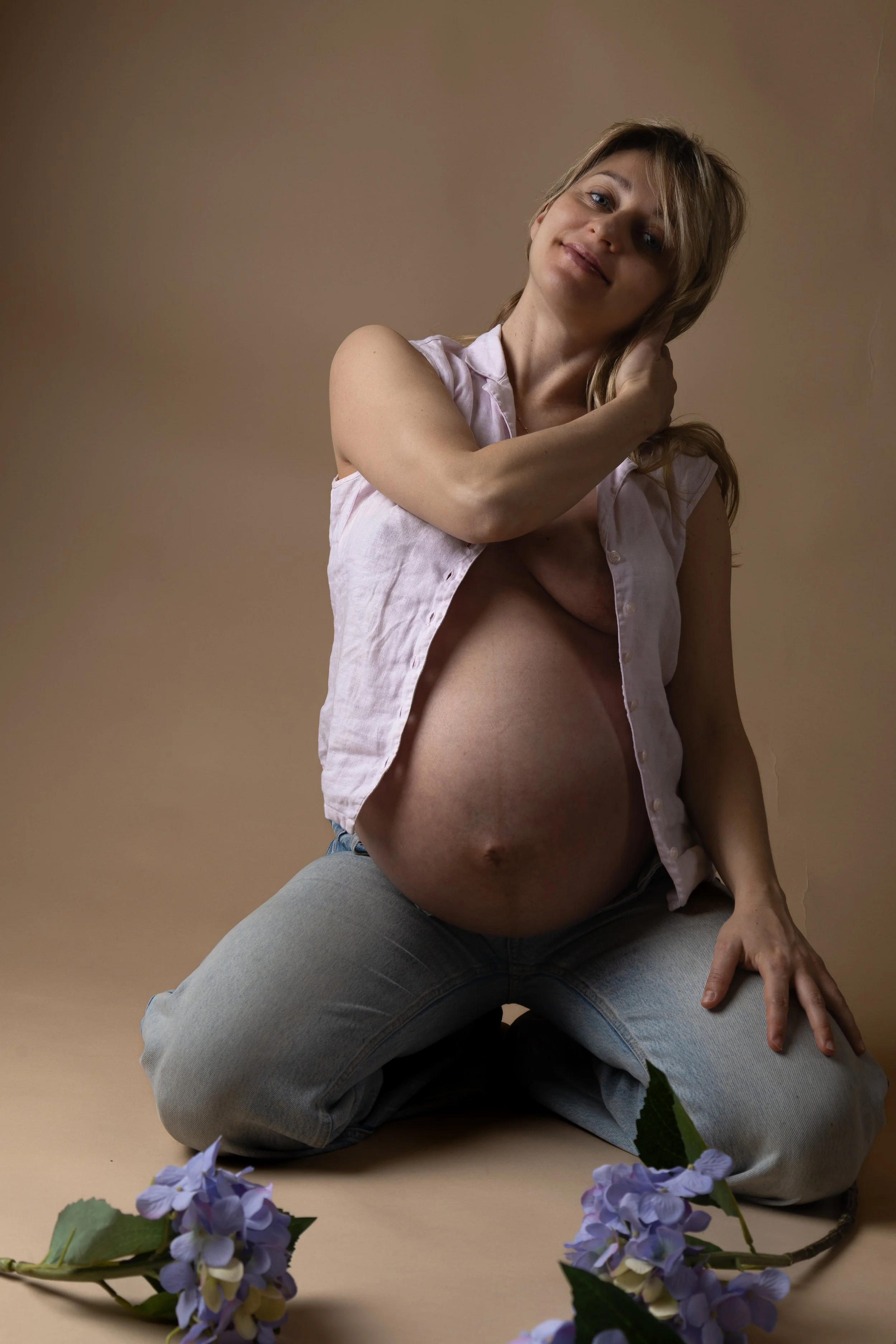 A pregnant woman with light skin and blond hair kneeling on the floor, wearing a partially unbuttoned pink shirt and blue jeans. She is smiling and touching her neck, with her other hand resting on her thigh. Purple hydrangea flowers are placed on th