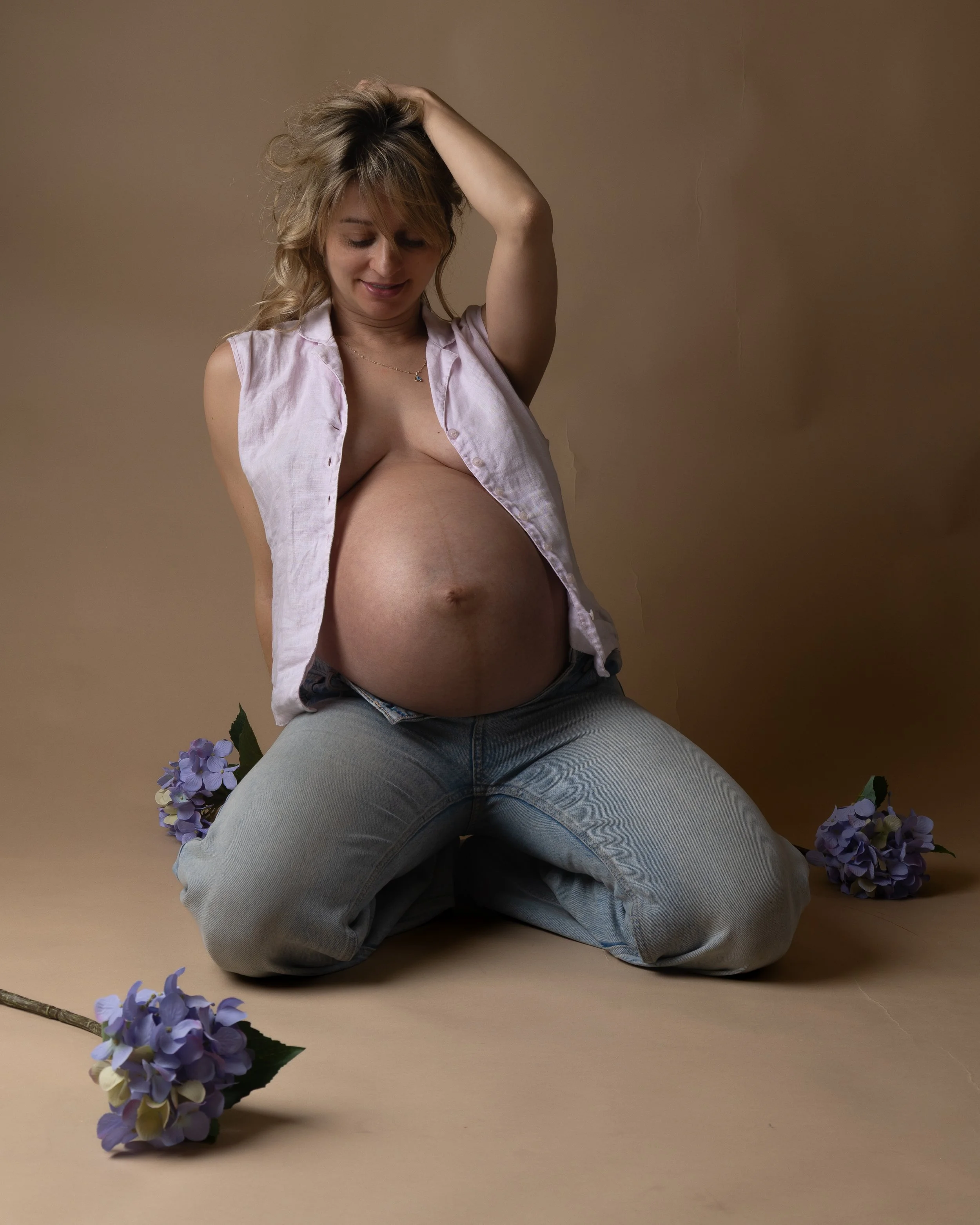 A pregnant woman kneeling on the floor with flowers around her, wearing a partially unbuttoned sleeveless shirt and jeans, smiling softly with her hand in her hair.