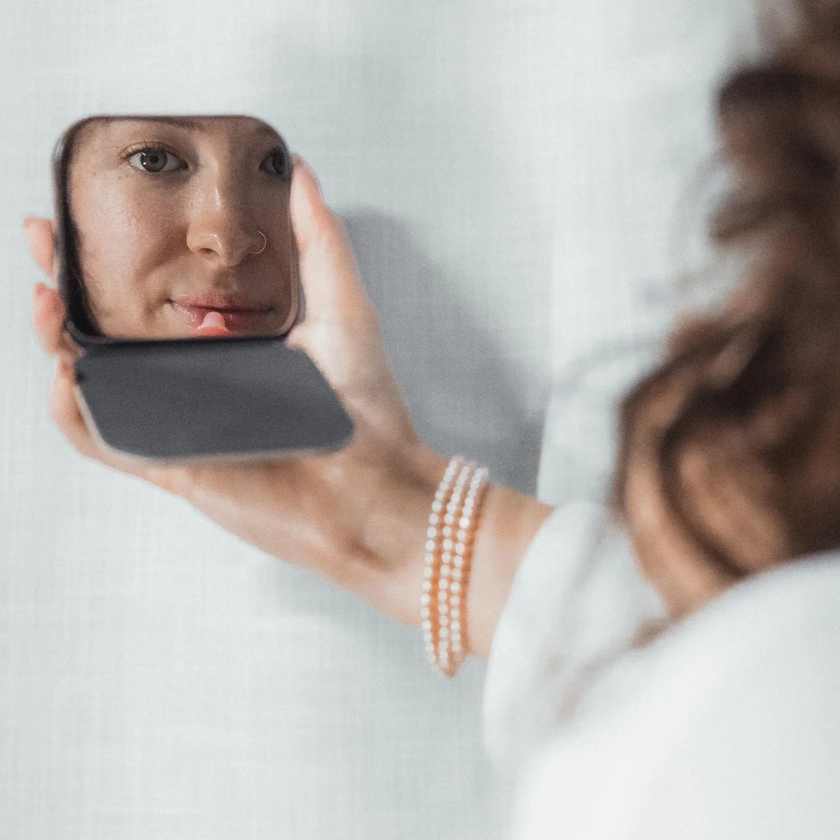 Person holding a small mirror reflecting their face with makeup, wearing a white shirt and a pearl bracelet.
