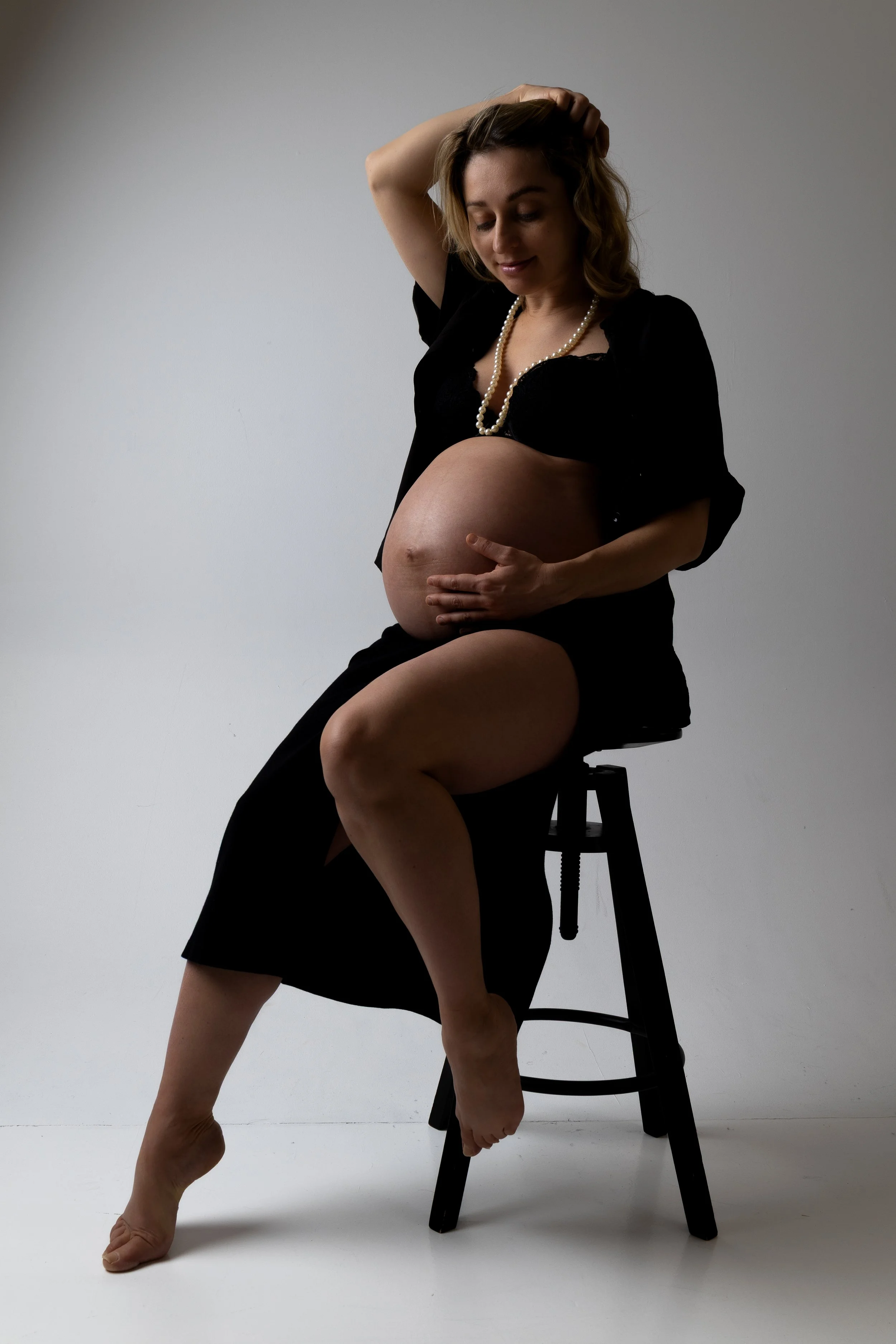 A pregnant woman sitting on a black stool in a studio setting, wearing a black dress and a pearl necklace, with one hand on her belly and the other touching her hair.