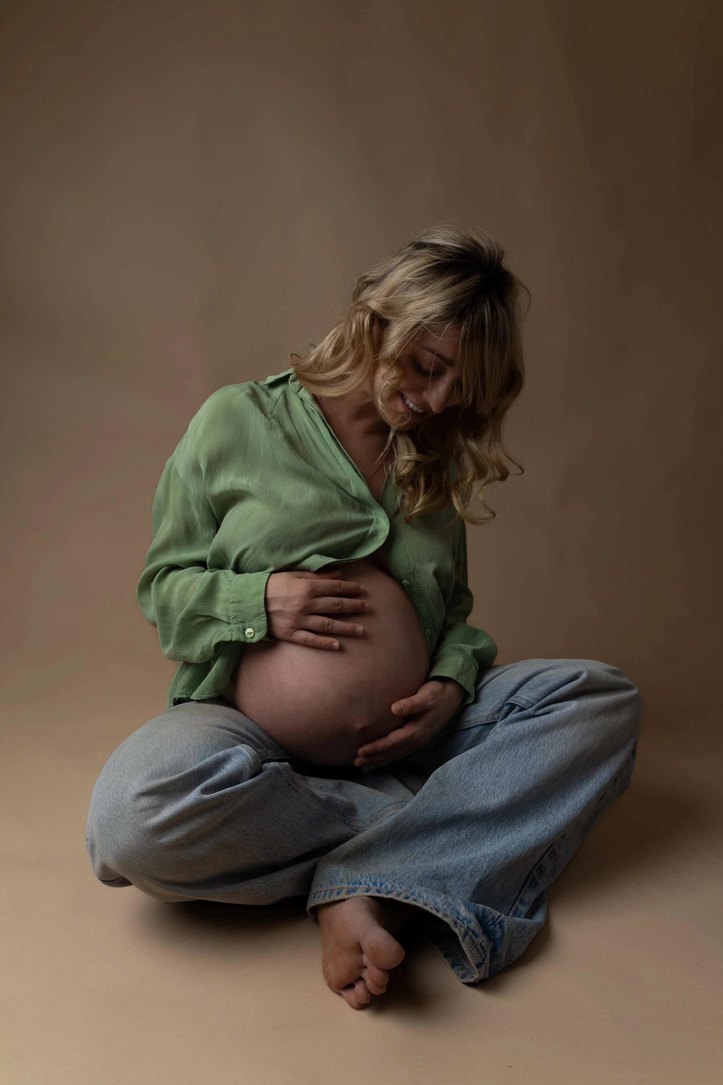 A pregnant woman sitting cross-legged on the floor, wearing a green blouse and jeans, smiling and looking down at her belly against a plain background.