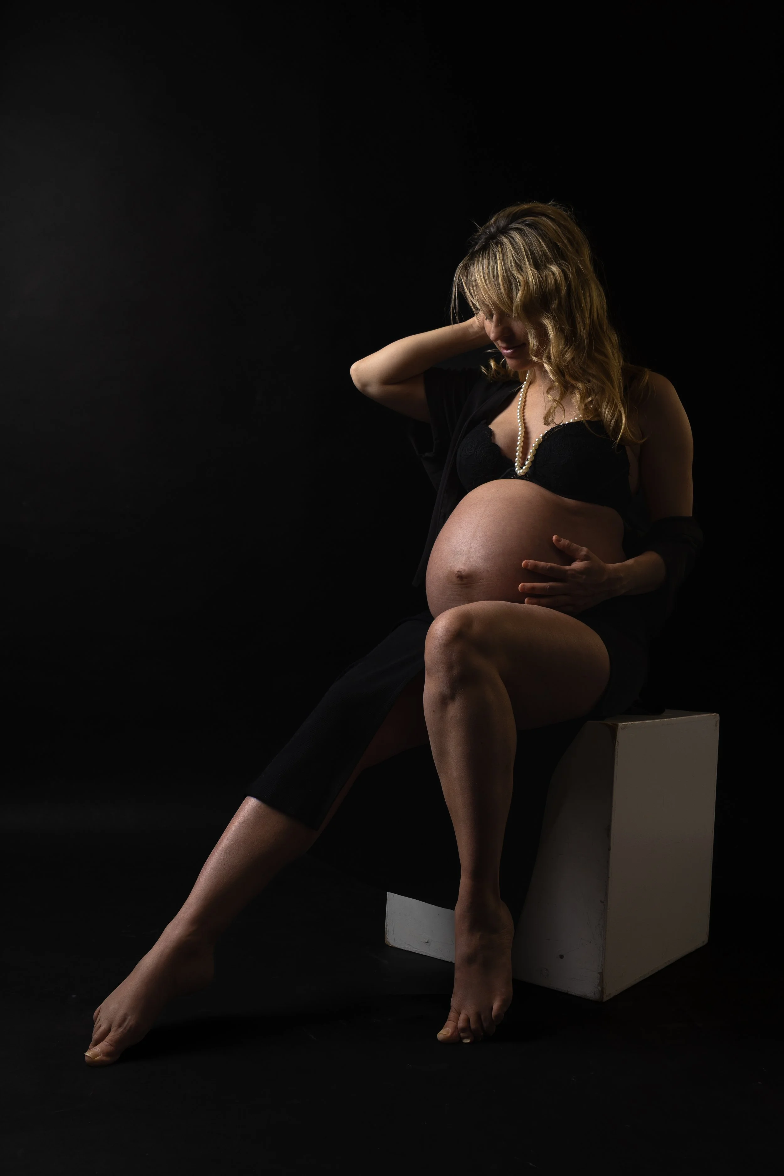 A pregnant woman sitting on a white block against a dark background, gently holding her belly, wearing black clothing and a pearl necklace.