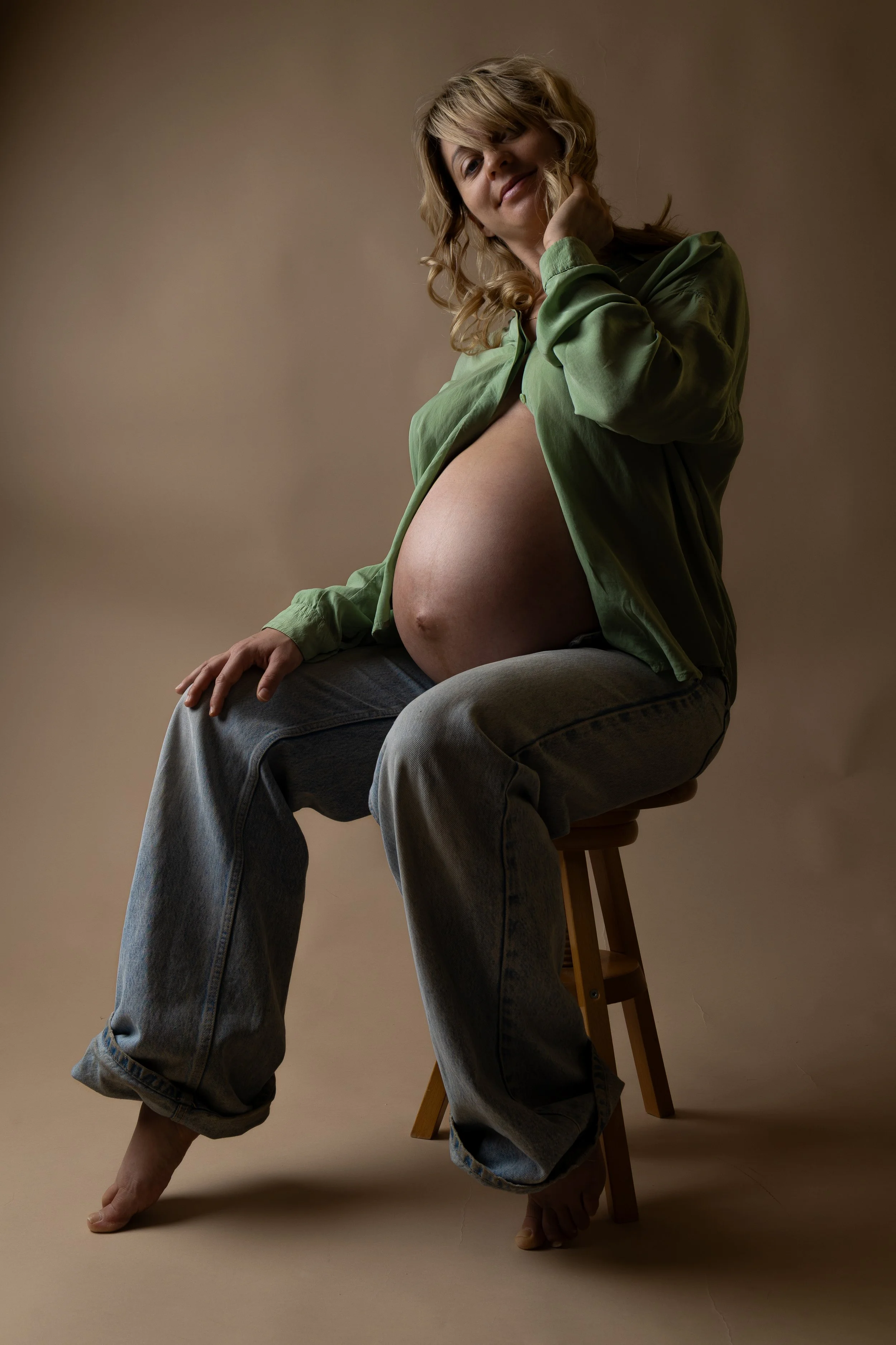A pregnant woman with blonde wavy hair, wearing a green shirt and gray jeans, seated on a wooden stool on a beige background, smiling gently.