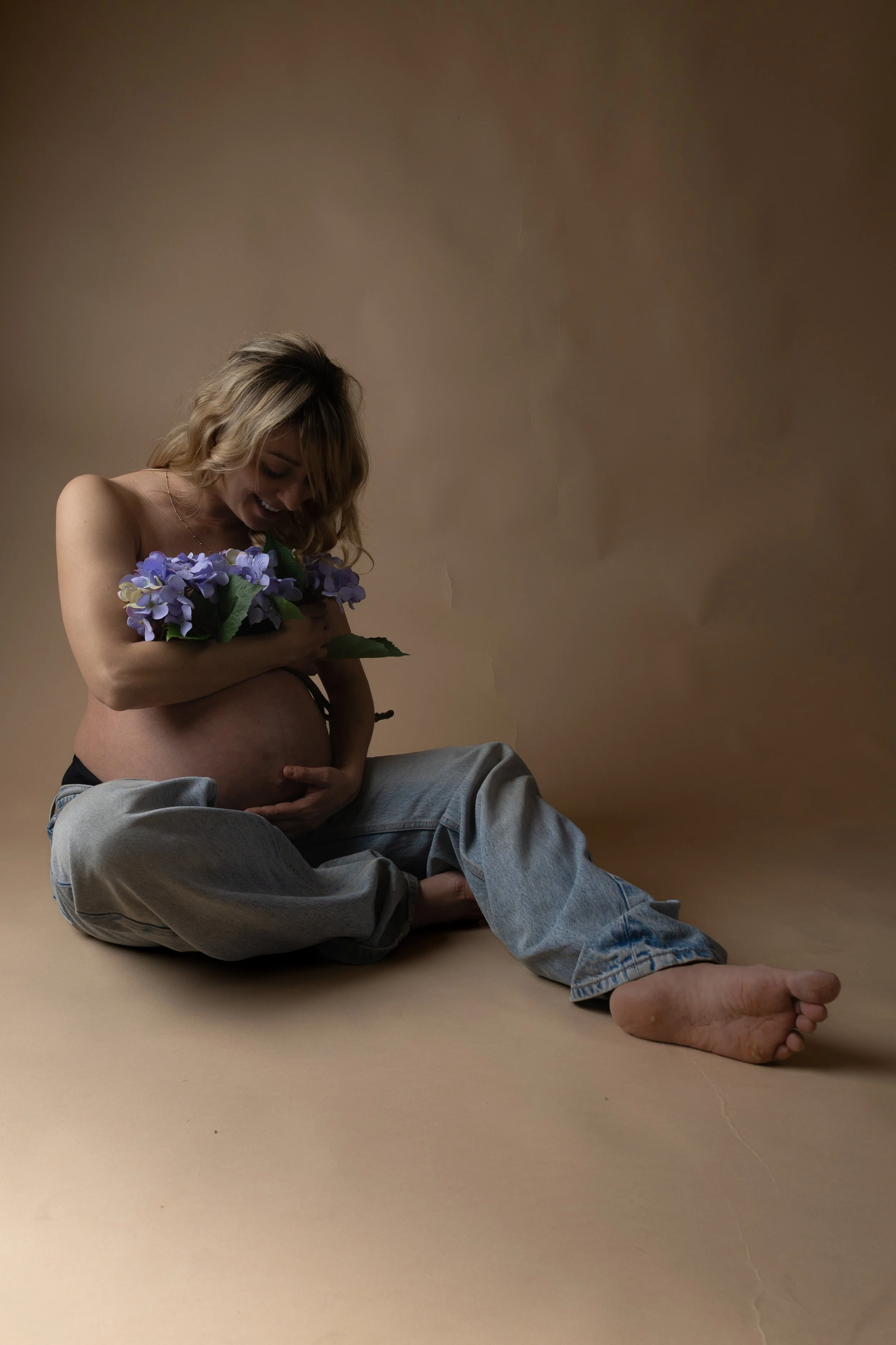 A pregnant woman sitting on the floor, holding purple flowers against her chest, looking down with a smile, wearing jeans and no top, against a plain beige background.