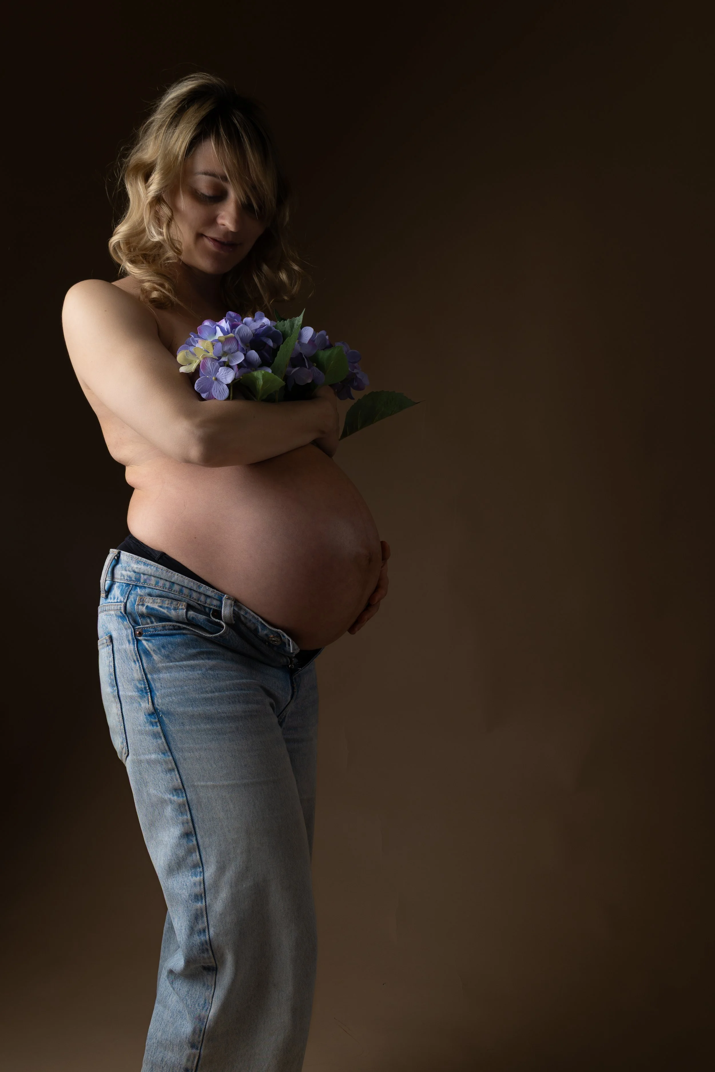 A pregnant woman with blonde hair, wearing jeans and holding a bouquet of purple and green flowers, standing against a dark background.