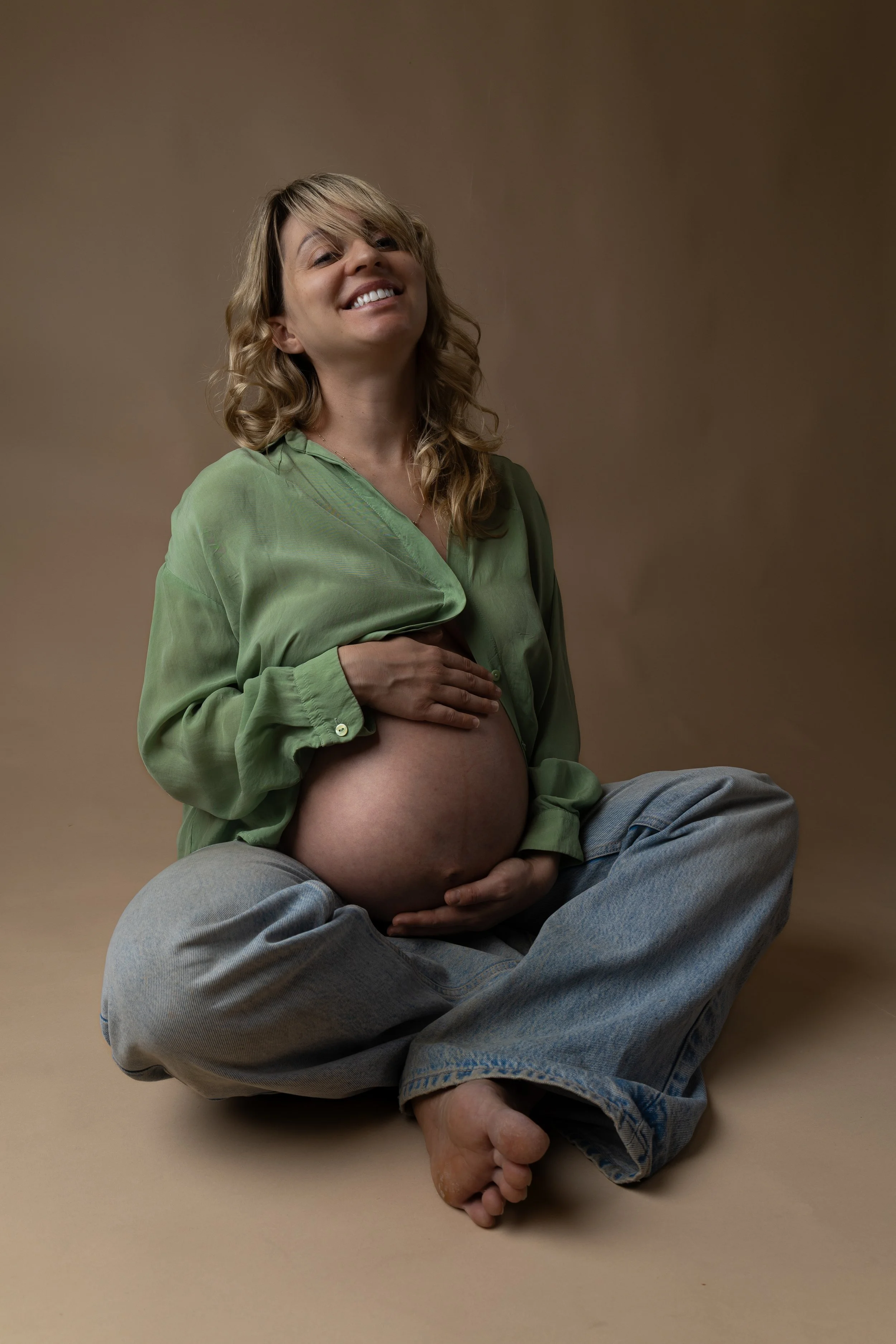A pregnant woman sitting cross-legged on the floor, smiling, with her hands resting on her belly, against a neutral background.