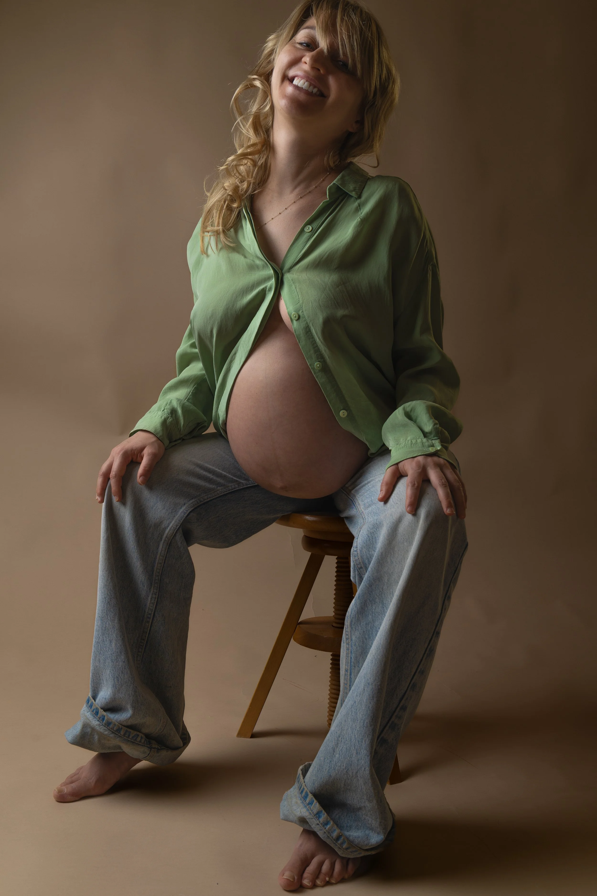 A pregnant woman with blonde hair, wearing a green silk shirt and jeans, sitting on a wooden stool against a plain background, smiling at the camera.