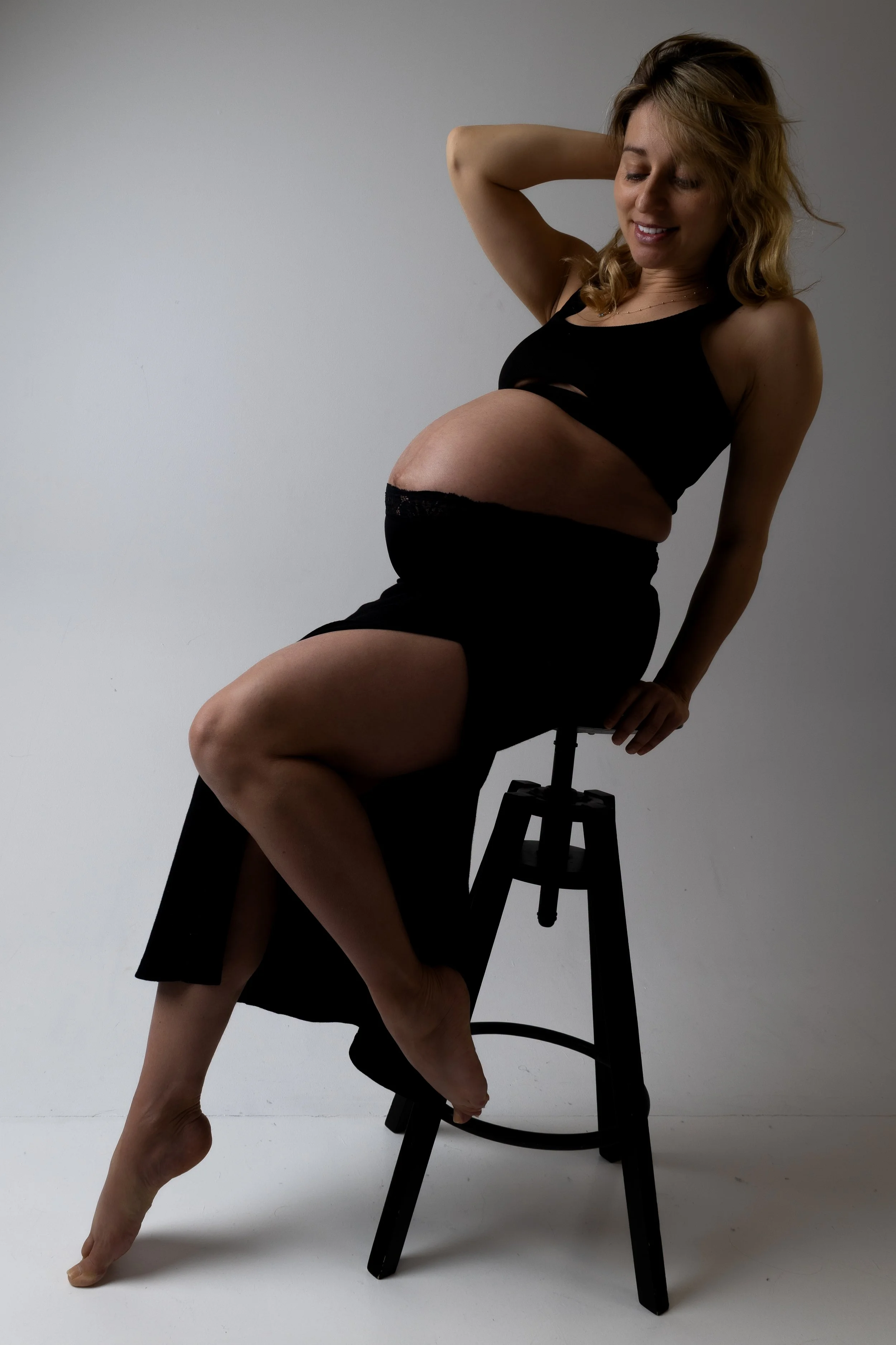Pregnant woman sitting on a black stool against a plain white background, smiling gently with one hand behind her head and the other resting on the stool, wearing a black tank top and skirt.