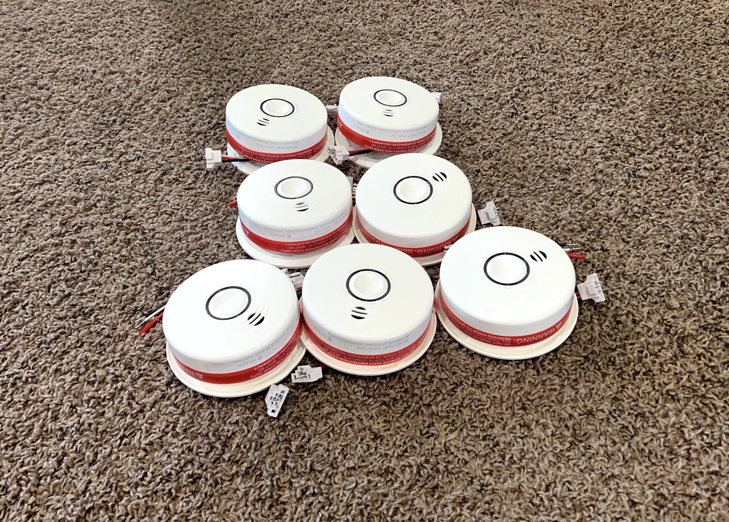 Six smoke detectors placed on a brown carpeted floor.