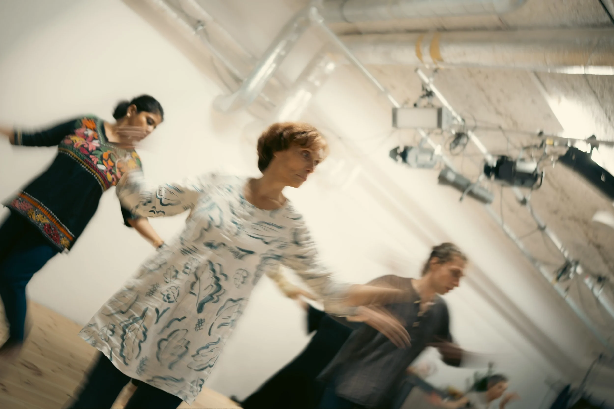 Multiple people dancing and holding hands in a dance class or social event, with studio lights and equipment visible in the background.