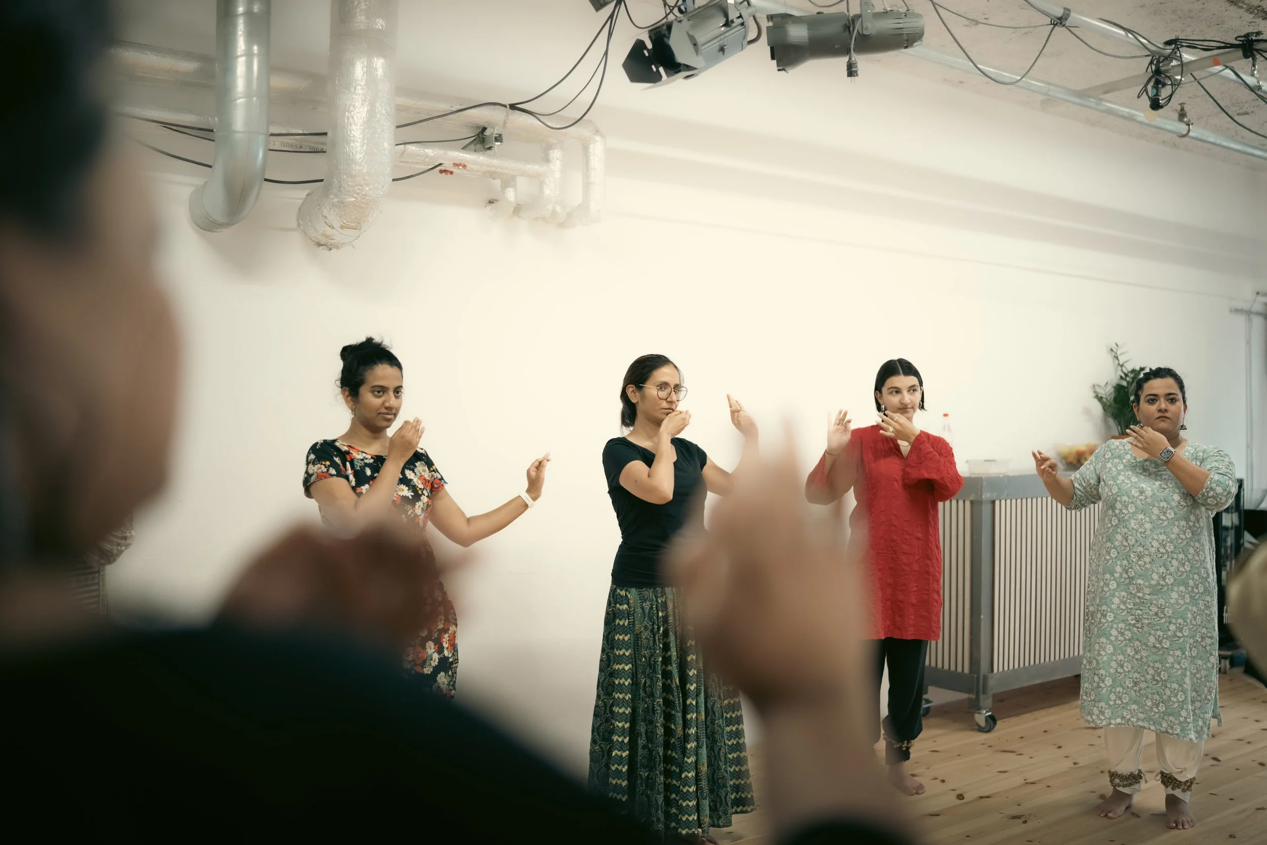 Group of four women standing in a line performing hand gestures, indoors with a white wall and ceiling pipes visible, all dressed in different colorful outfits.