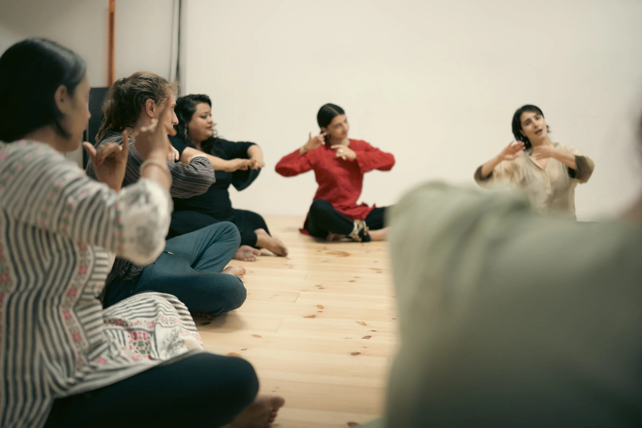 Group of women sitting cross-legged on wooden floor, participating in a discussion or activity, some making gestures or listening attentively in a room with plain white walls.