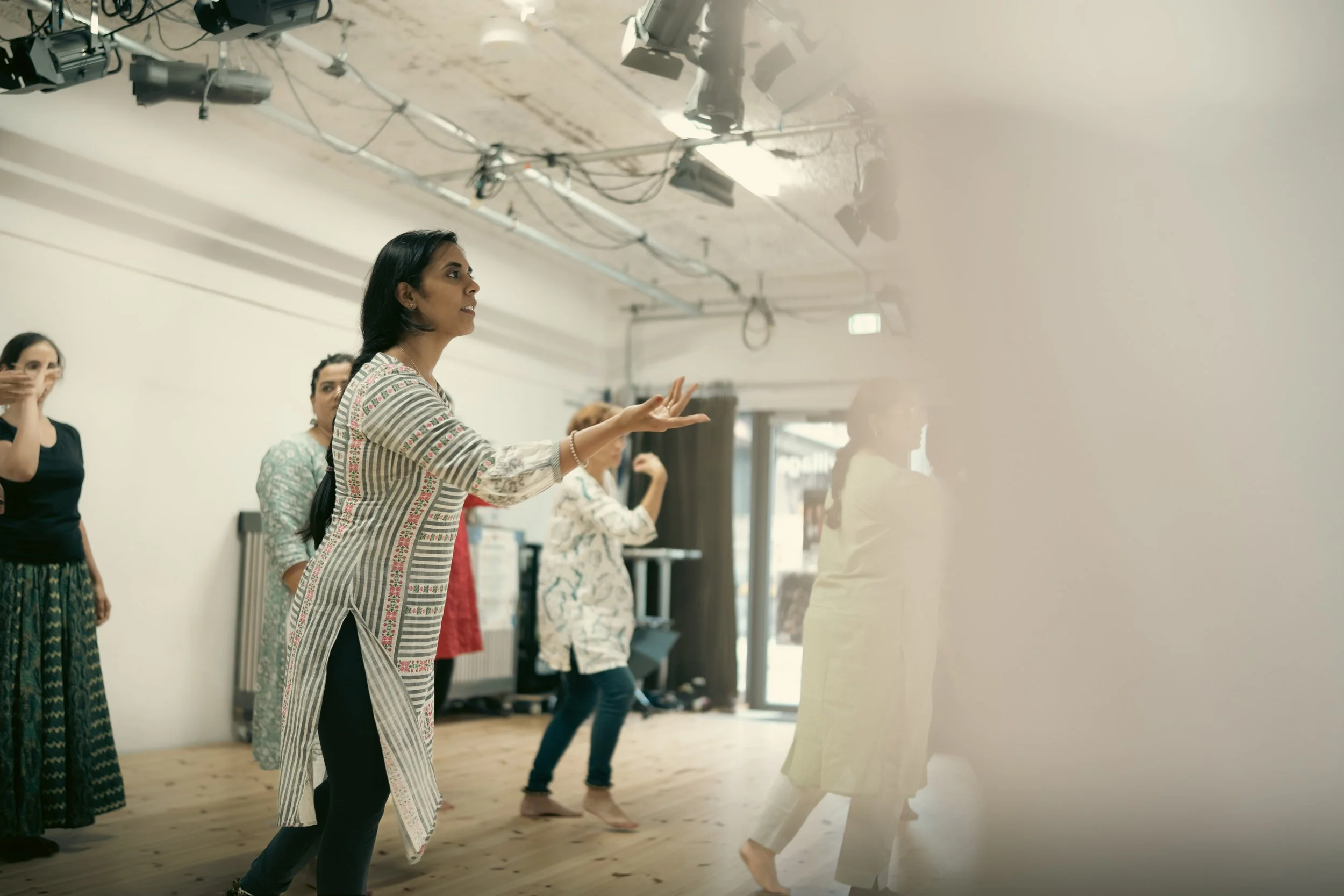 A group of women practicing dance moves in a dance studio. The women are in various positions and appear to be following a dance instructor. The studio has a wooden floor, a white wall, and a large mirror reflecting the women.