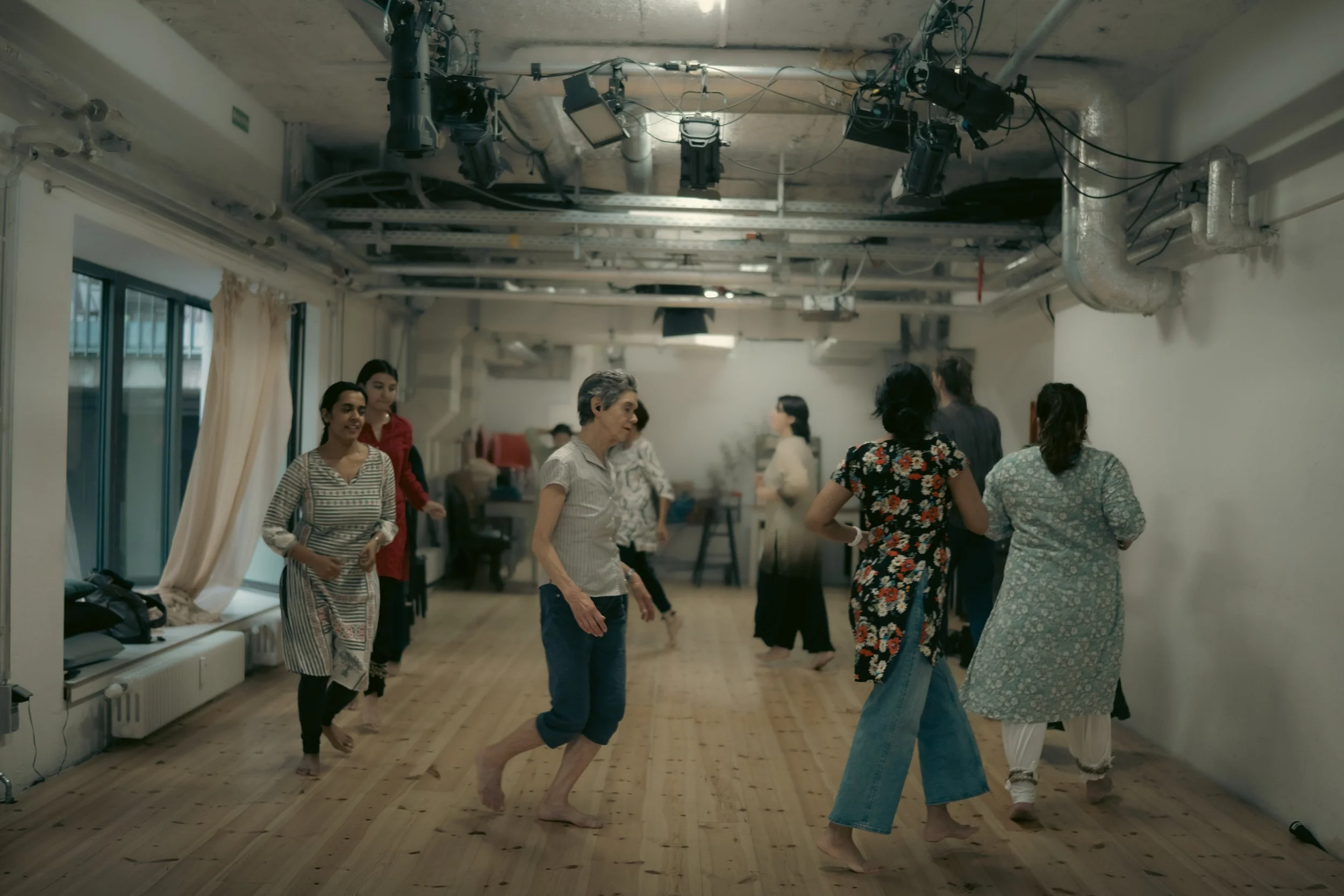 A group of people dancing and socializing in a room with wooden floors and a high ceiling, some with bare feet, while some are dressed in casual clothes with one person in a floral dress. Lighting equipment hangs from the ceiling.