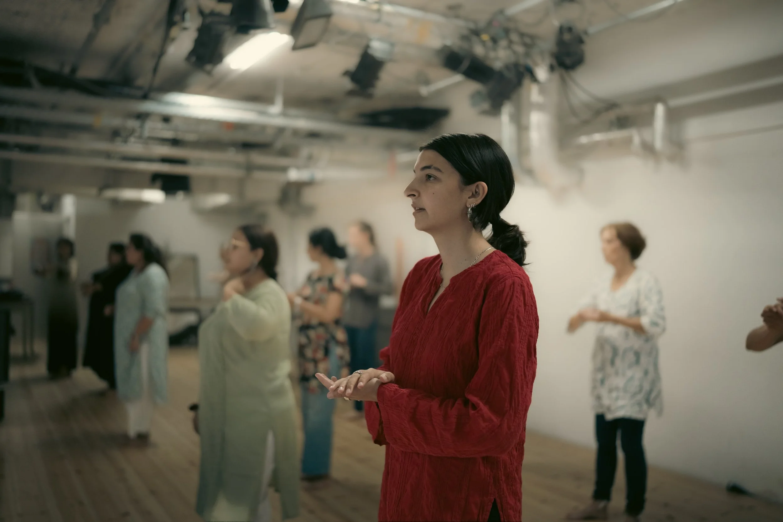 A woman in a red dress standing with her hands clasped in a large room with other women, some wearing light-colored clothing. The ceiling has exposed pipes and equipment, and the women appear to be participating in a group activity or gathering.
