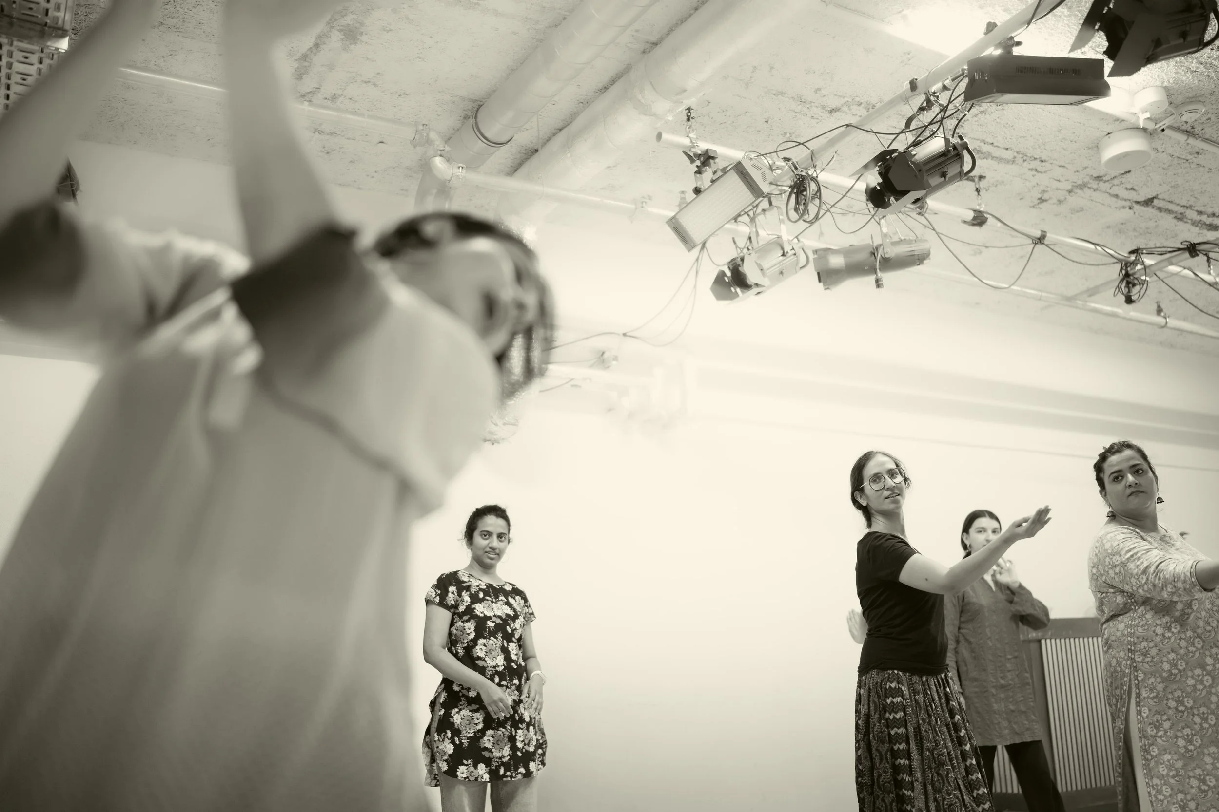 A group of women are standing in a room with their arms extended forward, engaged in a coordinated activity or exercise. Several electronic equipment and cables are hanging from the ceiling. The room has a white wall and exposed pipes on the ceiling.