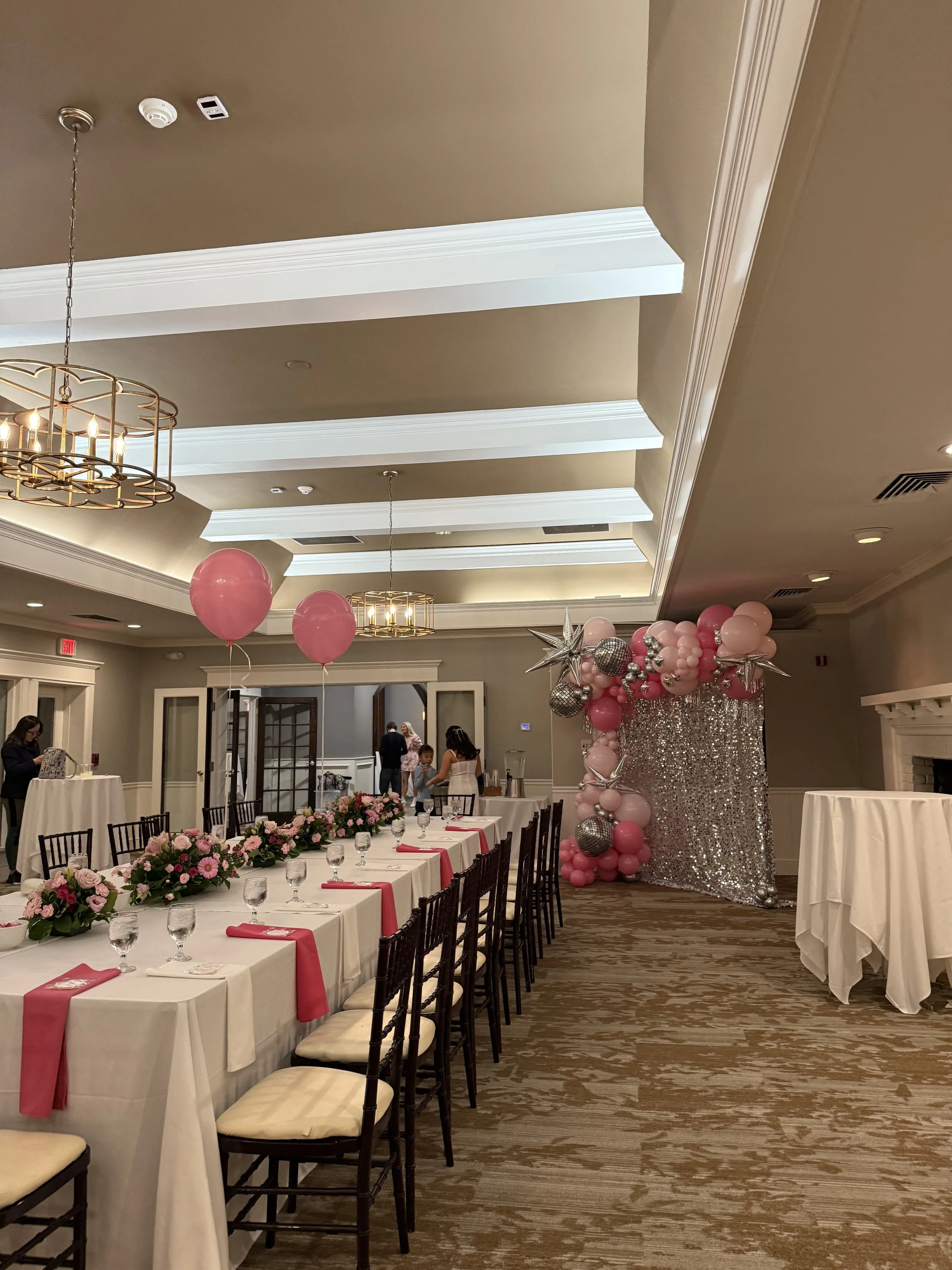 banquet room with long white table, black chairs; silver sequin pipe and drape with pink balloons attached