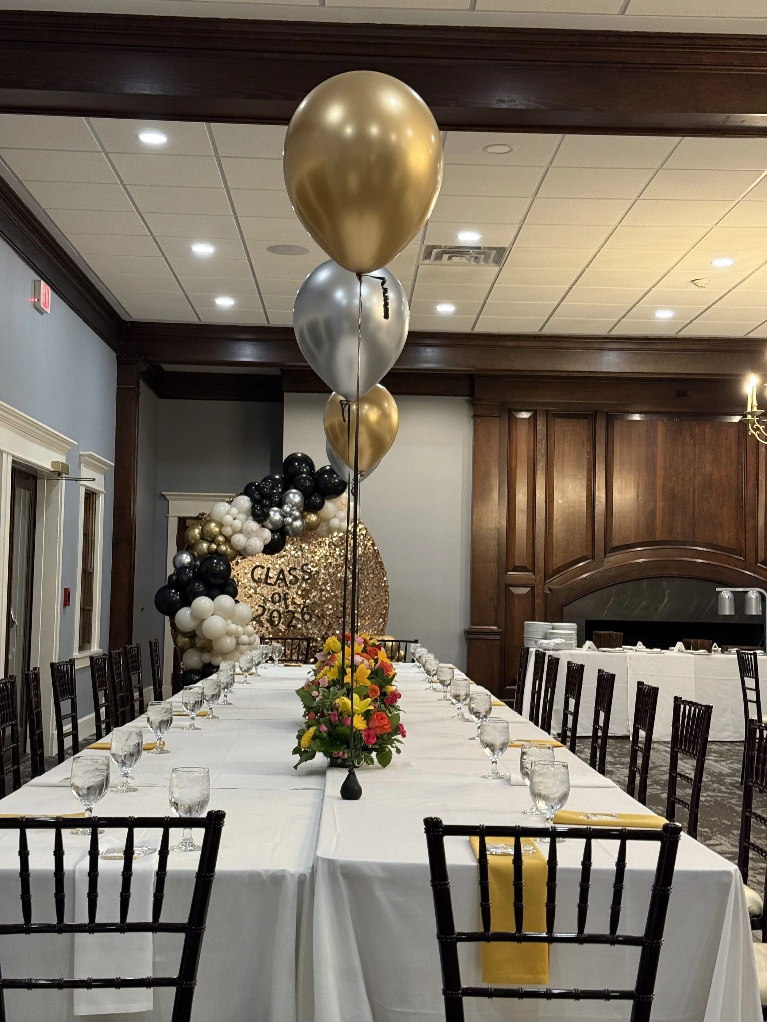 long white table with black chairs and yellow napkins; pink centerpieces and gold and silver helium balloons