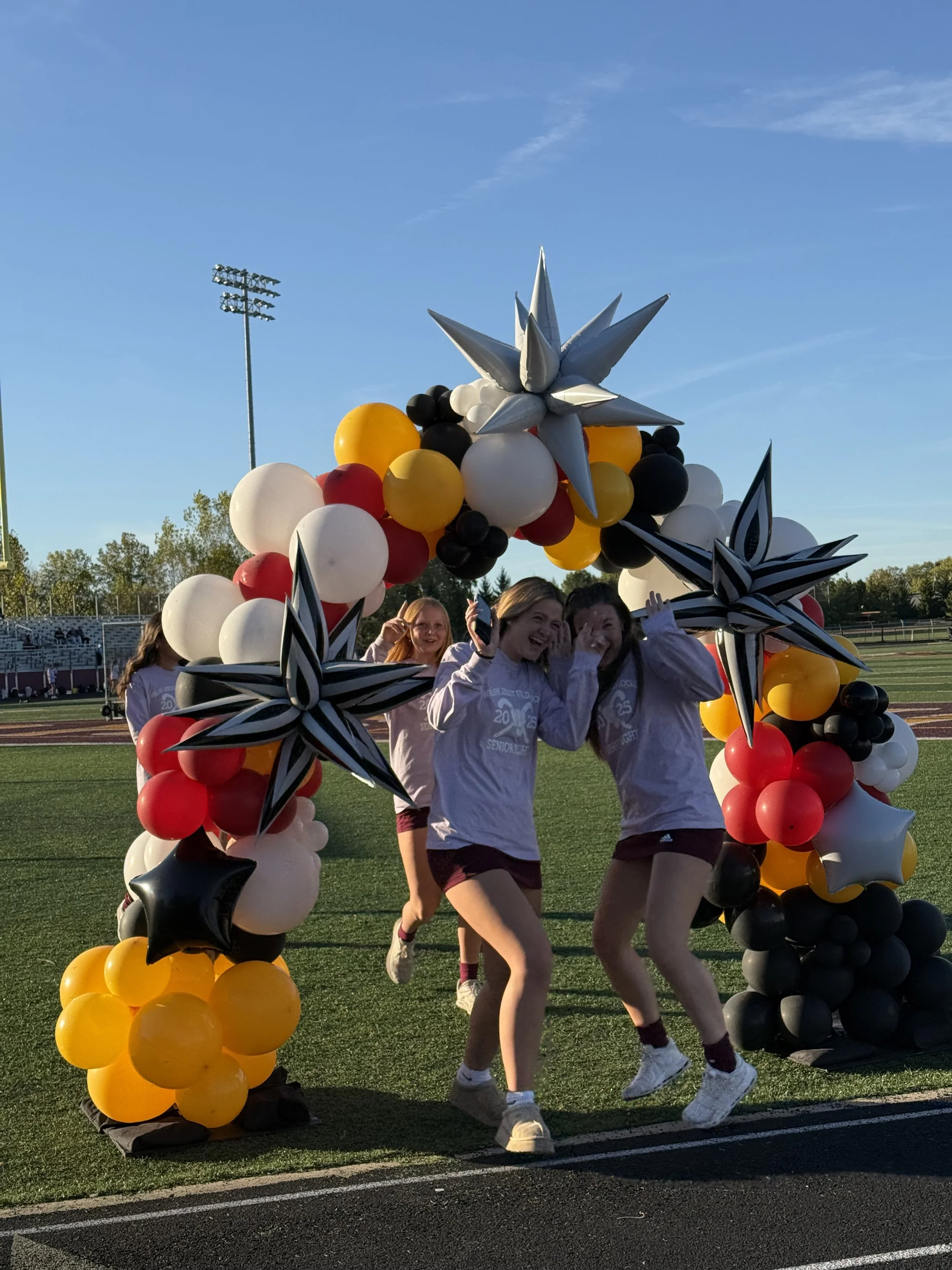 balloon arch outside on a football field with two teenage girls under the arch; the arch is made of yellow, black, white, and red balloons