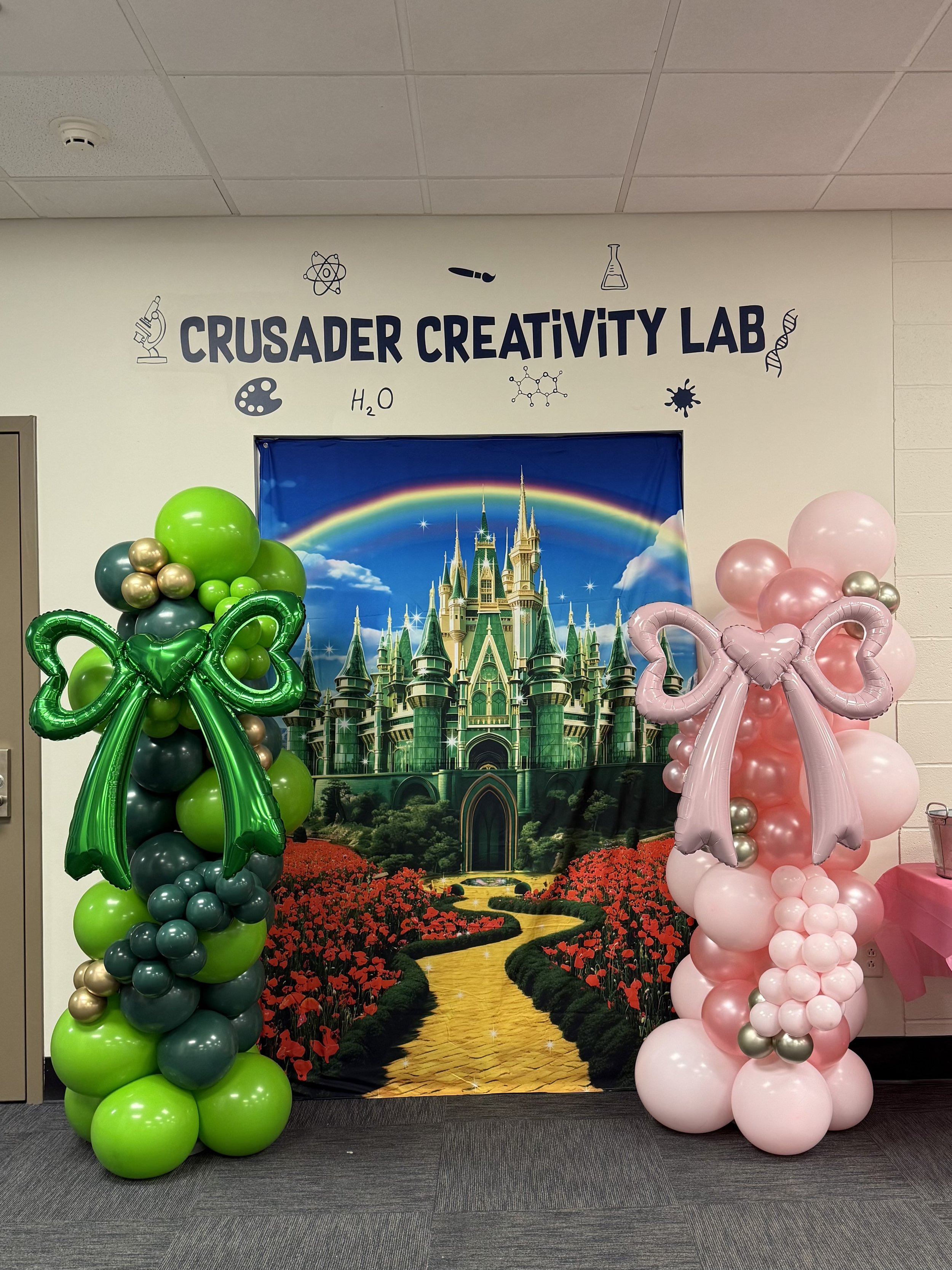 one green balloon column and white pink balloon column; columns are flanking a cloth backdrop with a Wicked movie theme