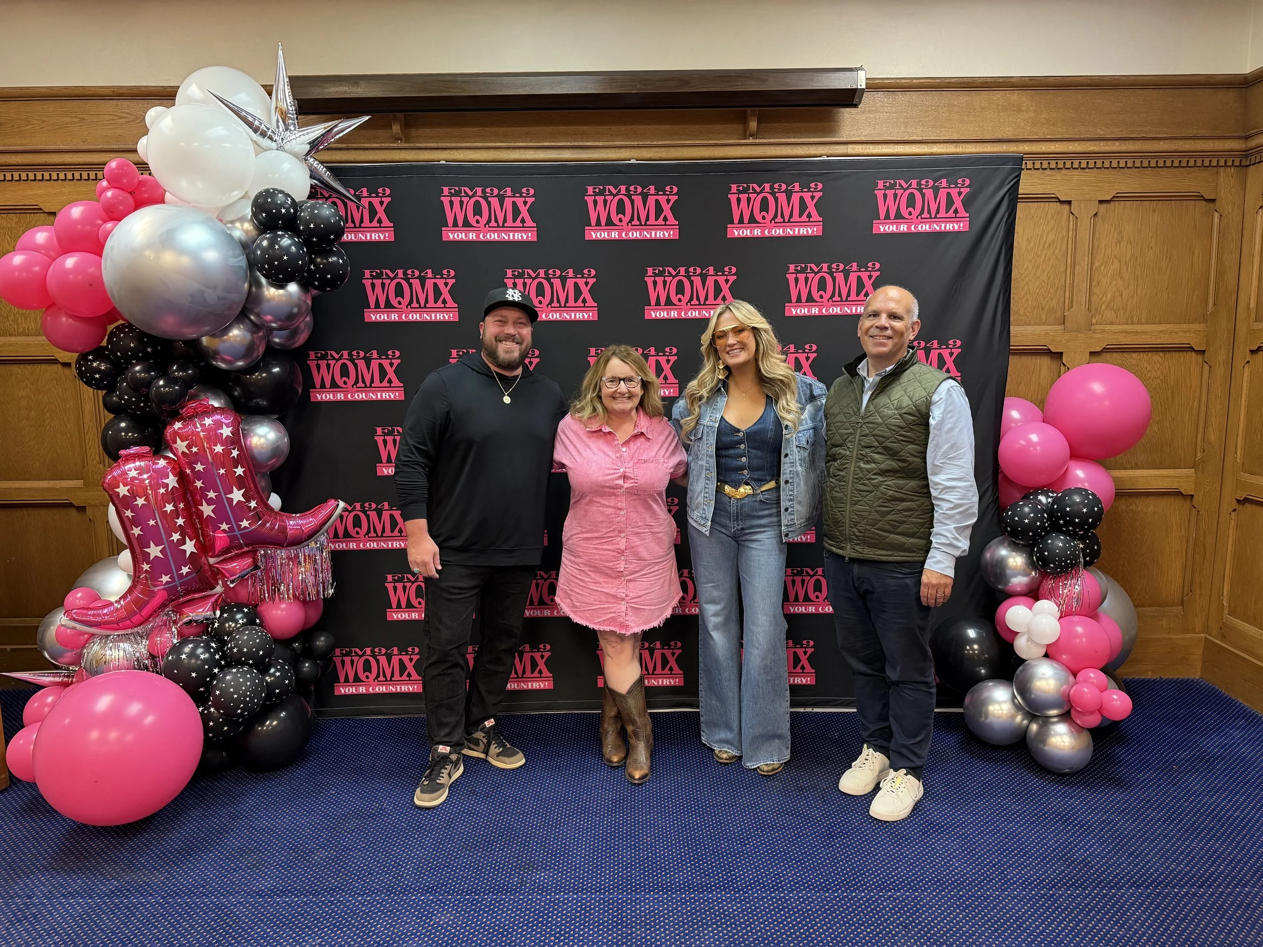 four people standing in front of a step and repeat for the radio station WQMX; pink, white, silver and black balloons are attached
