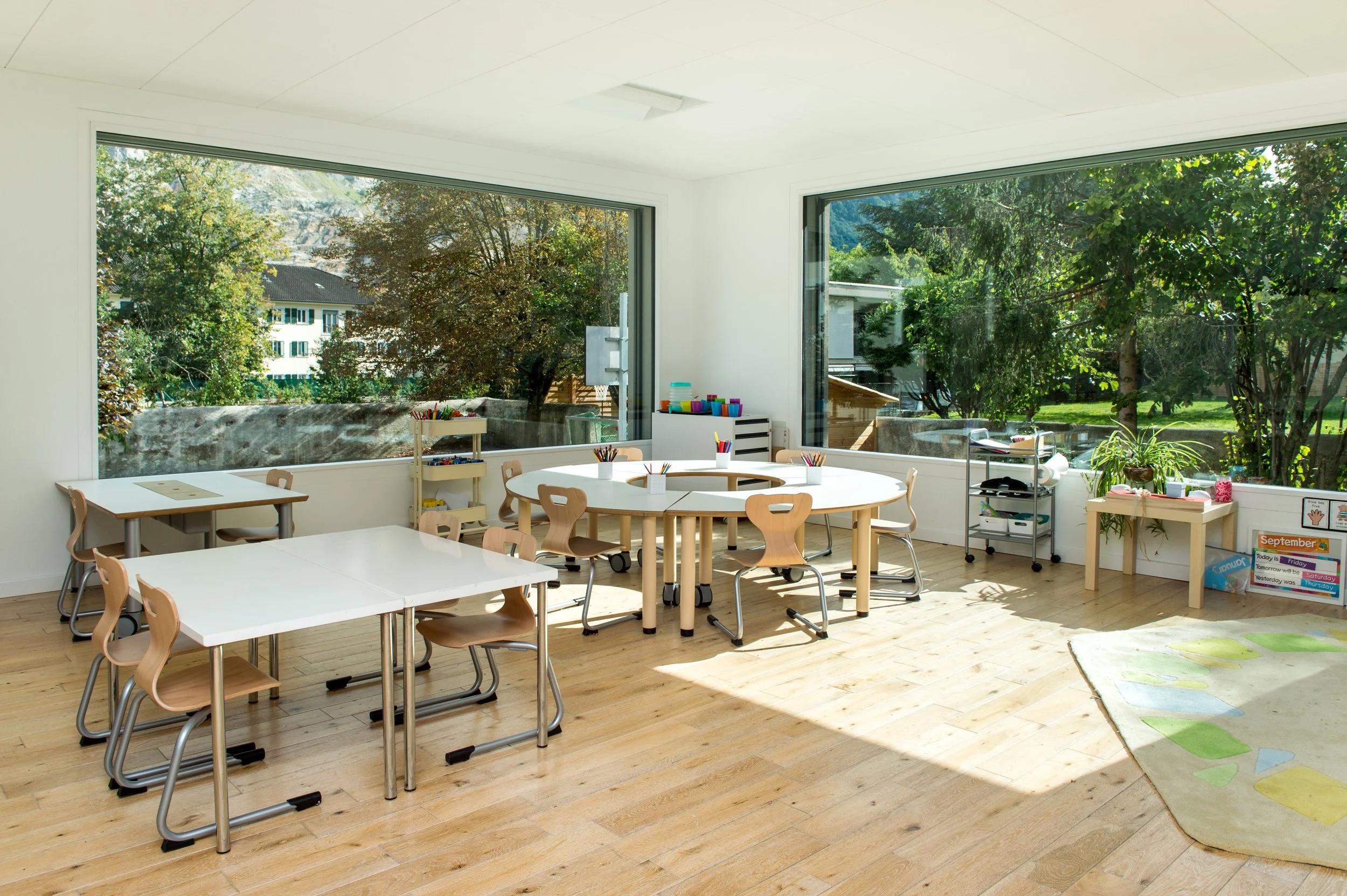 Salle de classe lumineuse de l’École Eden à Veyrier, Genève, avec fenêtres panoramiques, tables et chaises en bois, matériel pédagogique et environnement d’apprentissage accueillant.