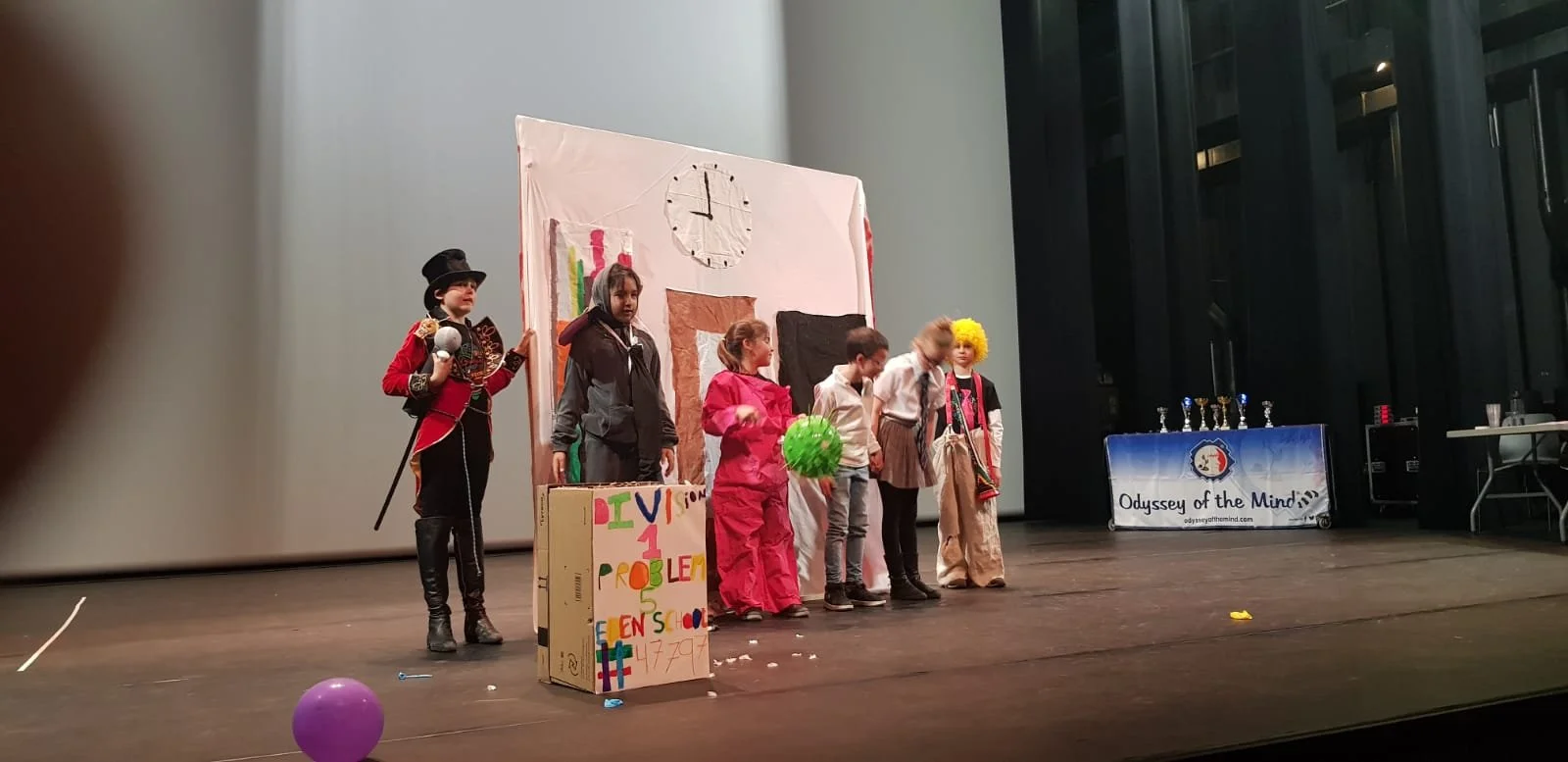 Groupe d'enfants en costume sur scène lors d'une pièce de théâtre scolaire, avec décor en papier, ballon vert et prix en trophées visibles à droite.