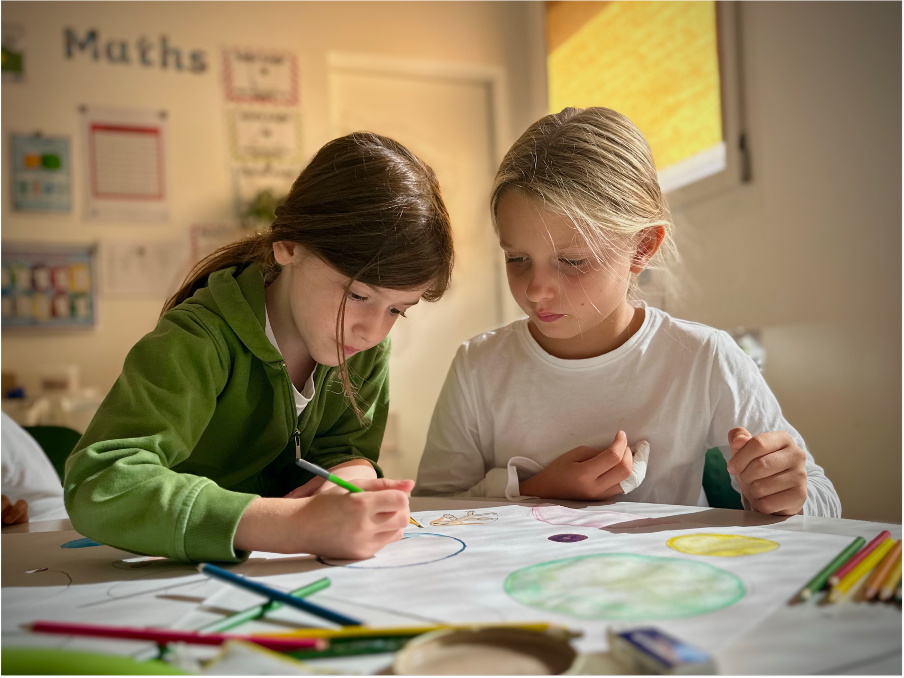 Deux jeunes filles à l’École Eden jouent avec des crayons de couleur devant une grande feuille de papier dans une salle de classe.