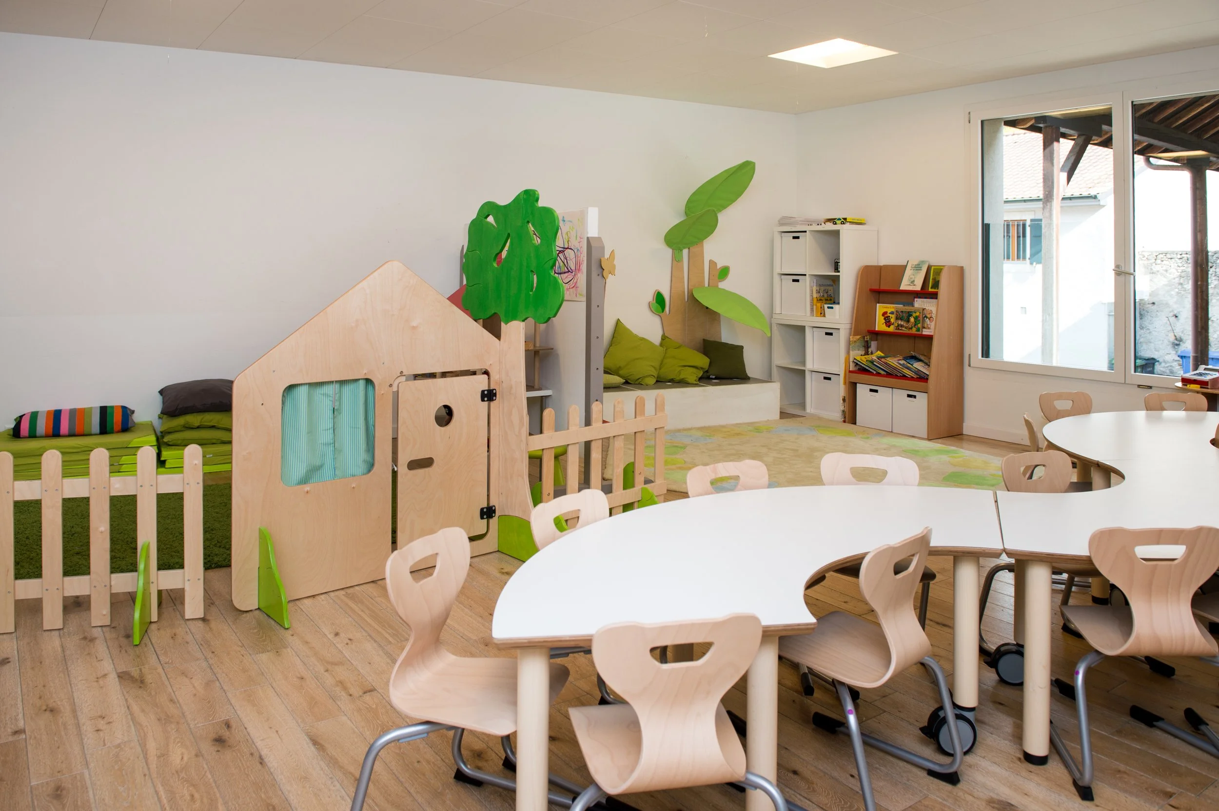 Salle de jeux pour enfants avec meubles en bois, tapis vert, murs blancs, grande fenêtre, petits sièges en bois autour de tables, espace de lecture et de jeu avec décorations en forme de feuilles et d'arbres.