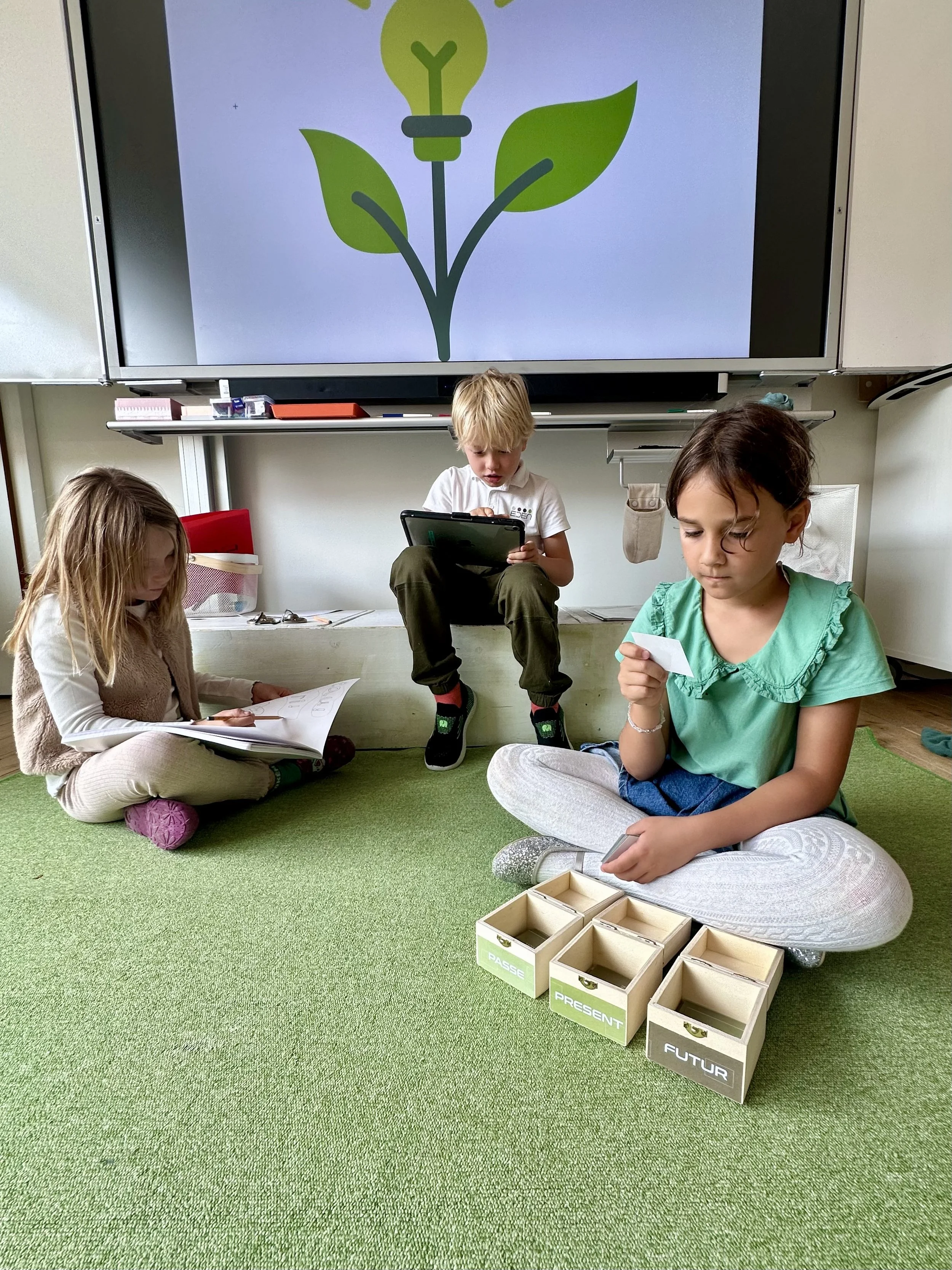 Three students at Ecole Eden in Veyrier, Geneva, engaged in a classroom activity using tablets, reading, and an interactive whiteboard, promoting modern learning and active participation.