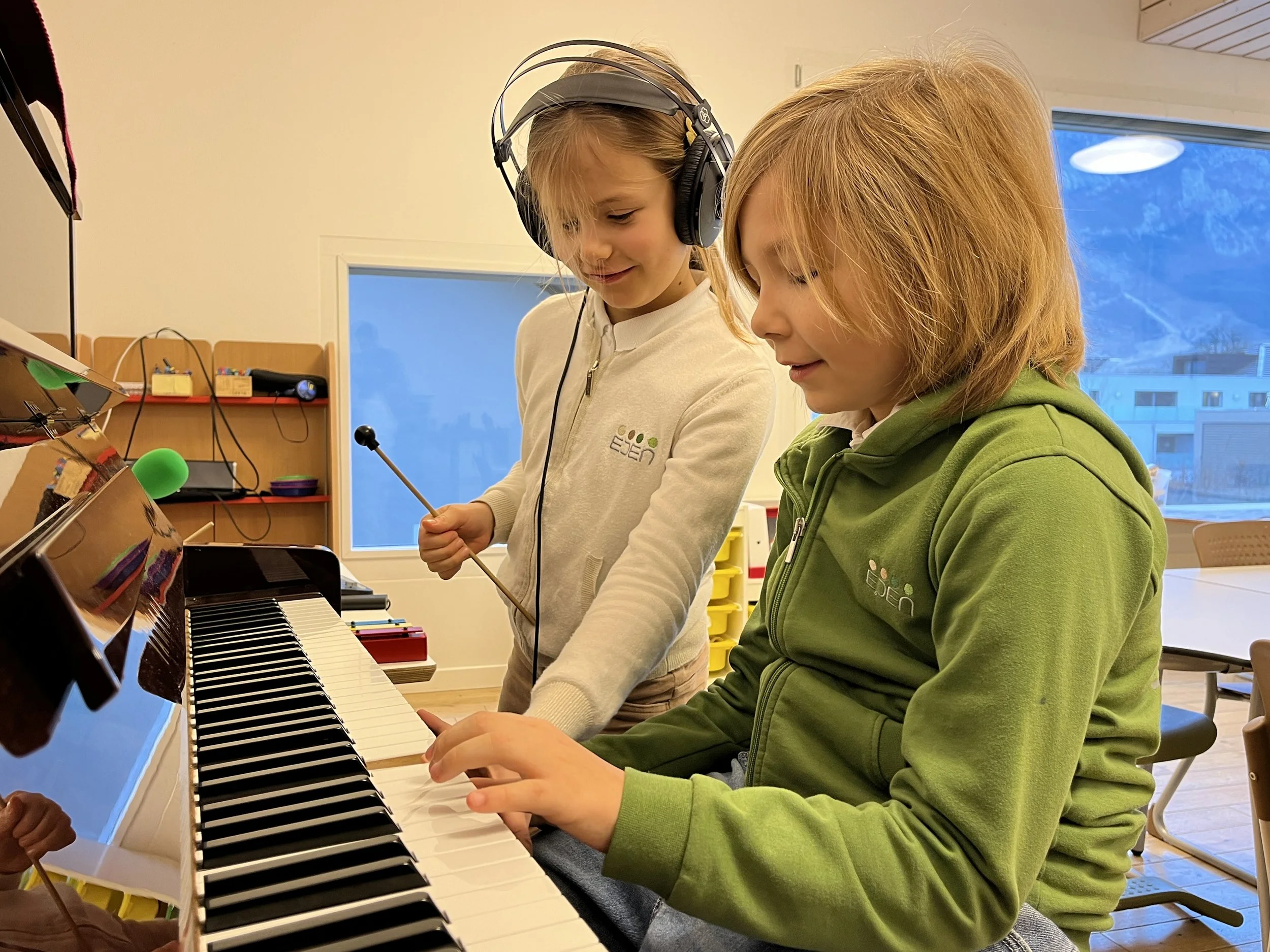 Deux jeunes filles jouent du piano, l'une avec des écouteurs et tambourin, dans une salle de musique ou d'apprentissage.