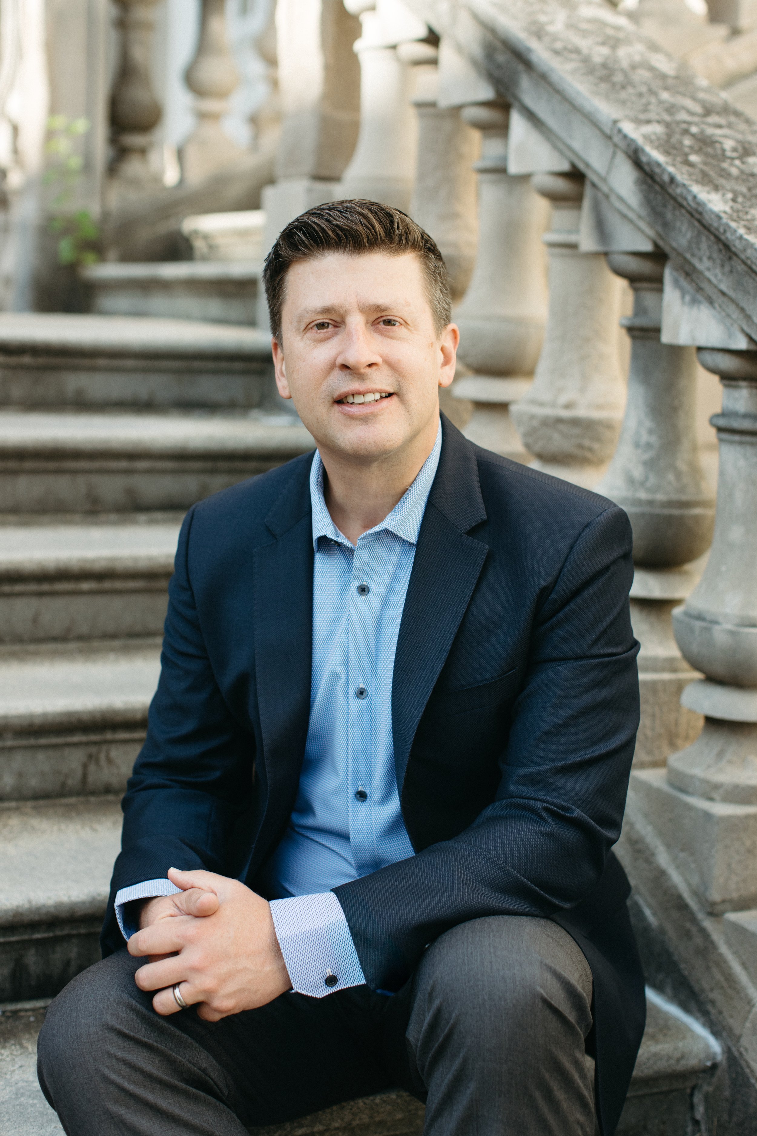 Headshot of Dan Heath in a dark suit and light blue shirt sitting on stone steps in front of a stone balustrade, with his hands clasped and smiling at the camera.
