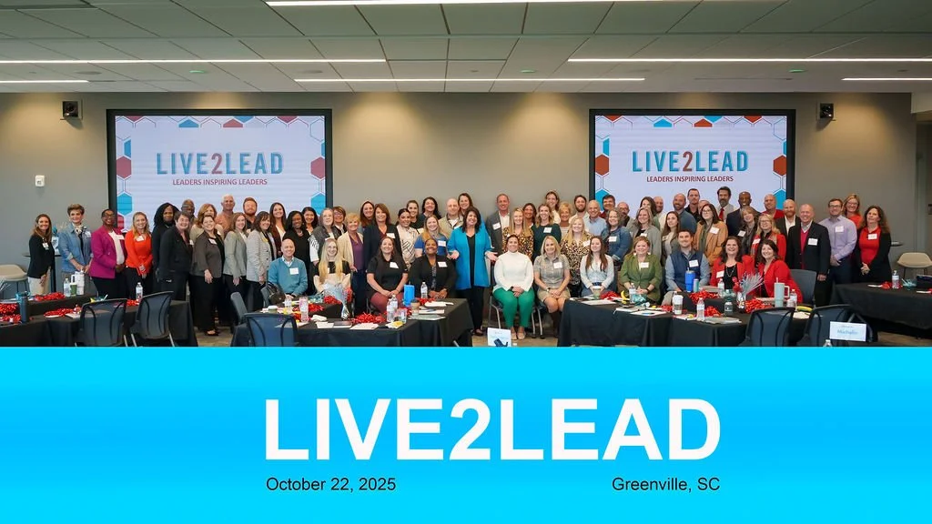 Group photo of conference attendees at the Live2Lead event in Greenville, SC, October 22, 2025, with two large screens displaying the event's name and tagline behind them.