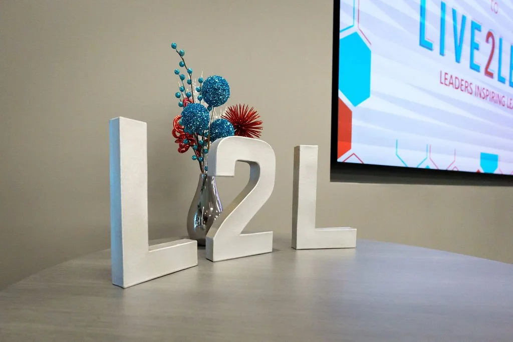White decorative letters spelling out 'L2L' arranged on a light-colored surface, with a silver vase holding blue, red, and teal decorative flowers. A presentation screen displays 'LIVE2025' with partial text and graphics in the background.