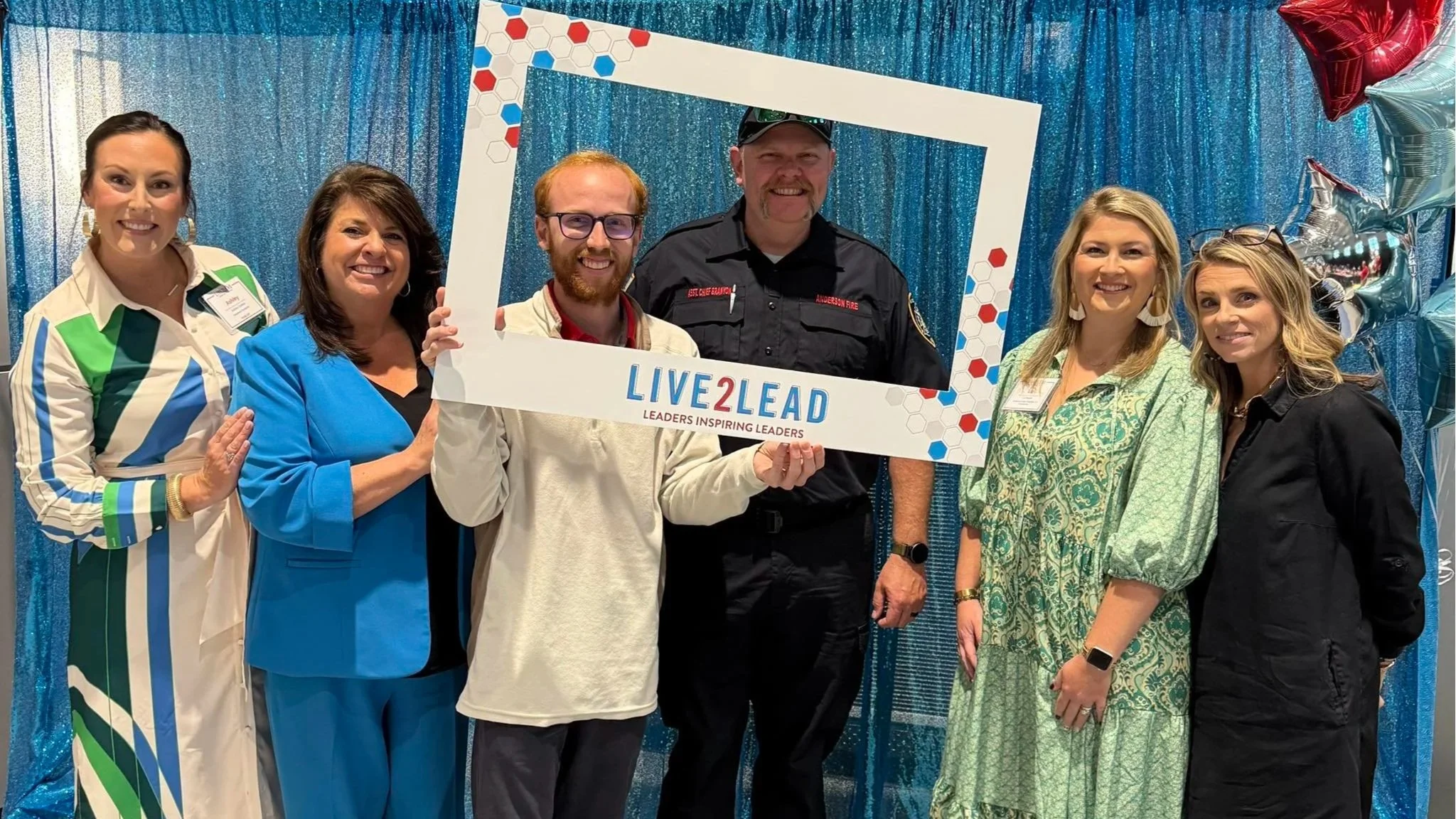 Group of six people, four women and two men, standing in front of a blue sparkly backdrop and balloons. One woman is wearing a striped dress, another in a bright blue outfit, a third in a green patterned dress, and the fourth in black. One man is wearing a beige jacket, and the other in a black firefighter uniform with a badge. They are smiling and holding a large white frame with the words 'LIVE2LEAD LEADERS INSPIRING LEADERS' at the bottom.