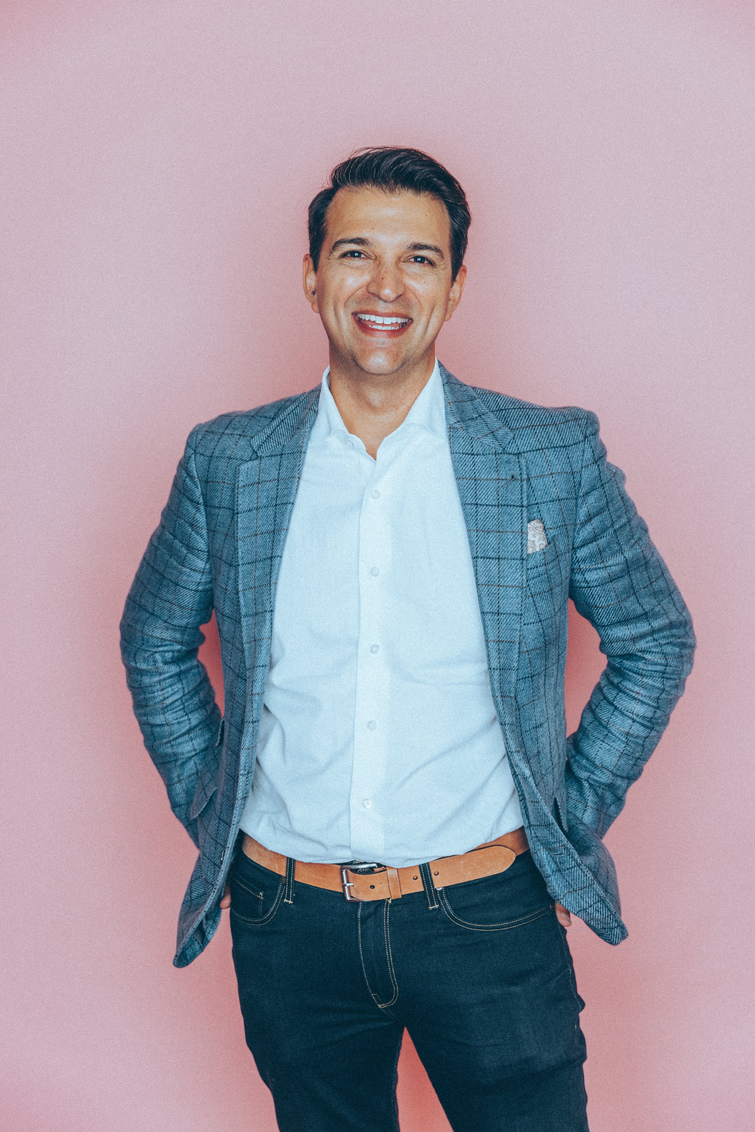 Headshot of Rory Vaden, smiling with hands on hips, wearing a gray plaid blazer, white shirt, dark jeans, and a tan belt, standing against a pink background.